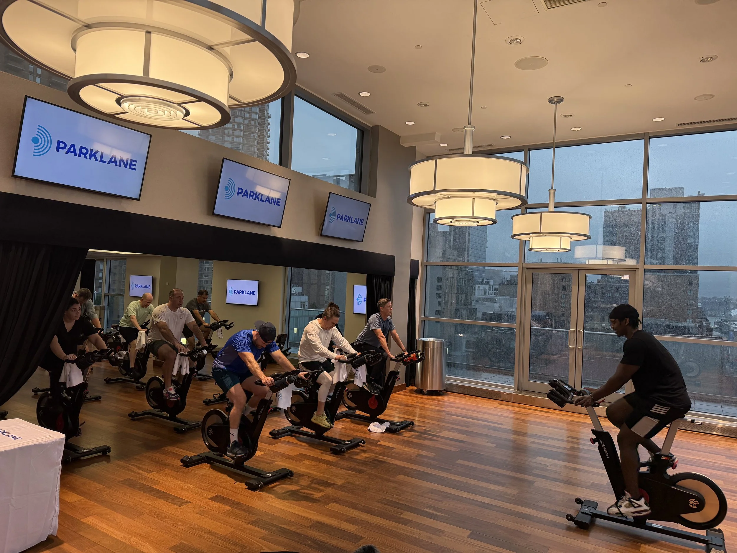 A group of people participating in a spinning class inside a modern gym, with large windows showing a cityscape, and multiple screens displaying the 'PARKLANE' logo.
