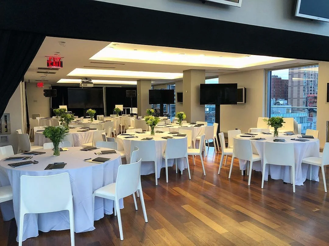 A bright conference room set up with round tables covered with white tablecloths, each decorated with green floral centerpieces, and surrounded by white chairs. The room has large windows and wall-mounted screens, with a cityscape view outside.