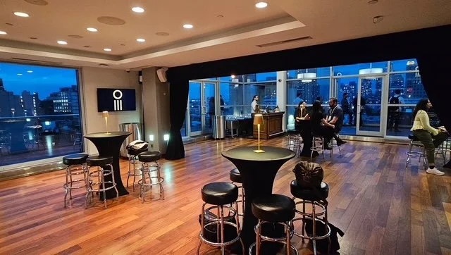 A modern indoor lounge with high tables and barstools, large windows showing cityscape at dusk, and some people socializing.
