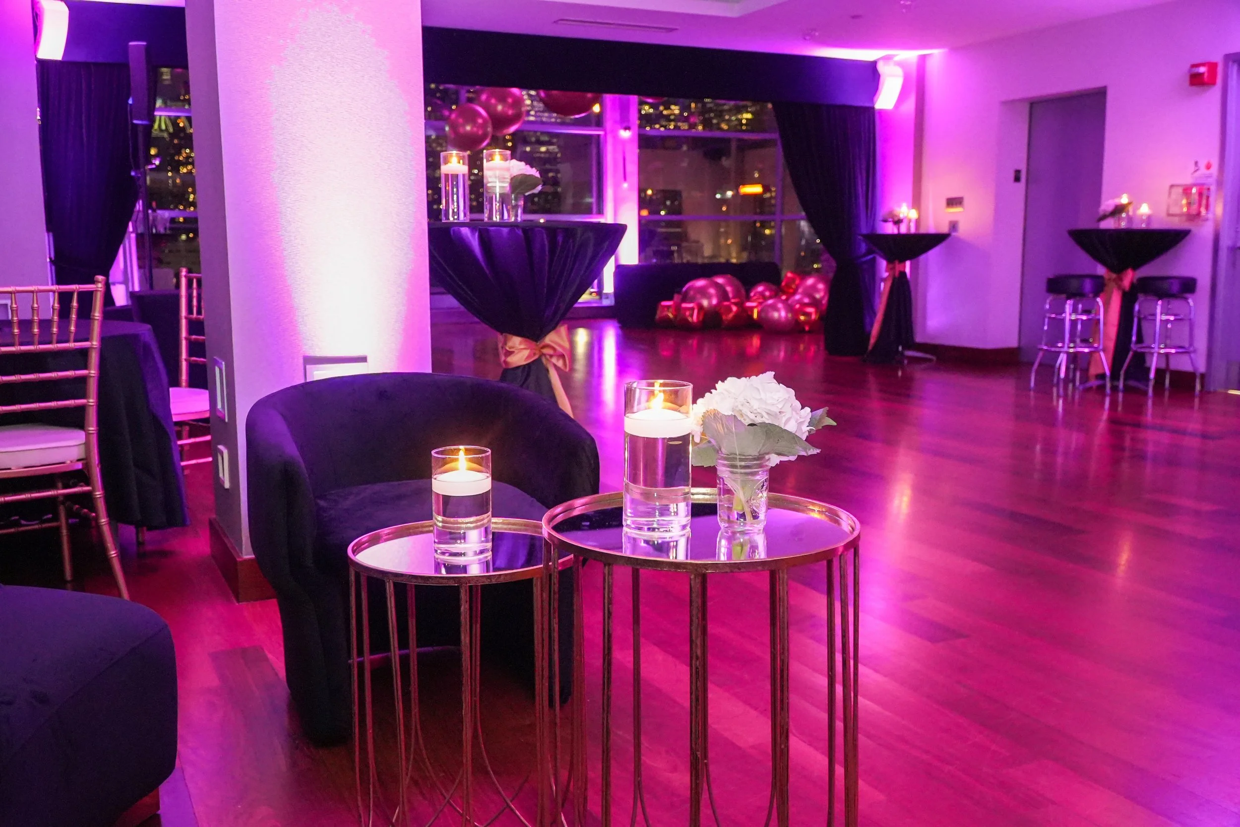 A dimly lit room with purple and pink lighting, decorated for a celebration, featuring balloons, lit candles, and floral arrangements.