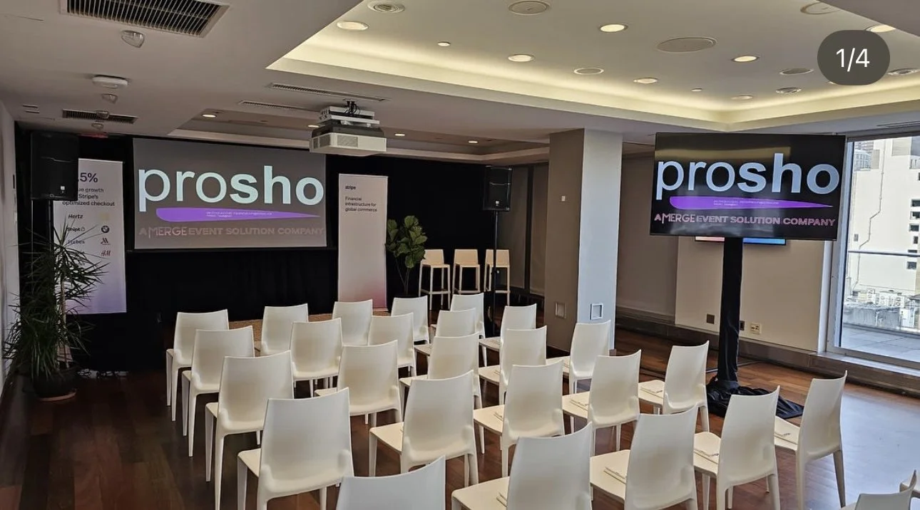 Conference room with white chairs arranged in rows, large screens displaying 'prosho' and event details, a black stage with speakers, and large windows showing a cityscape.