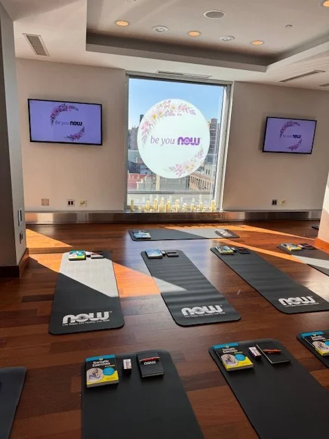 Yoga mats arranged on a hardwood floor in a bright room with large windows, TV screens on the walls displaying a logo, and an urban cityscape outside the window.