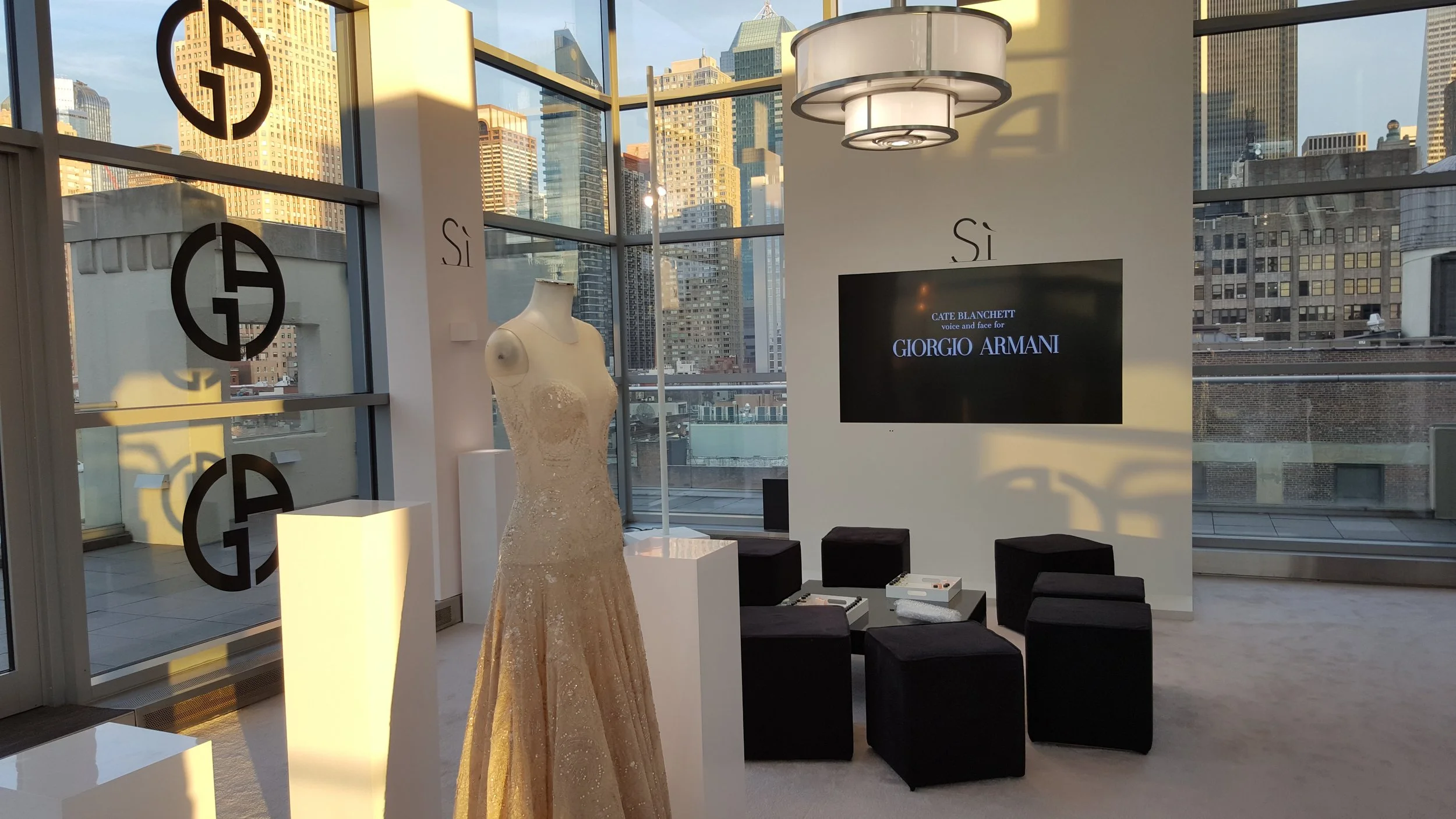 Interior of a modern gallery or display space with a mannequin dressed in a large, ornate gown, black stairs, digital screen with text, large windows with city skyline, and decorative lighting fixtures.