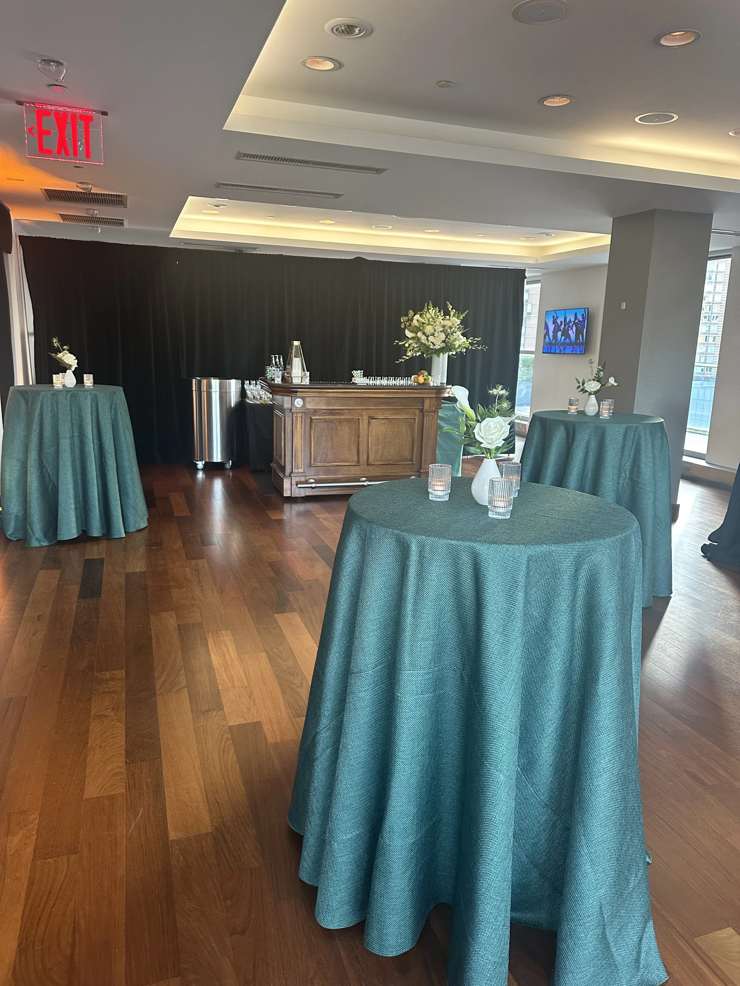 A room decorated for a social event with three round tables covered in teal tablecloths, each with vases of white flowers and small tealight candles. In the background, a wooden bar with glasses, bottles, and a large flower arrangement stands against