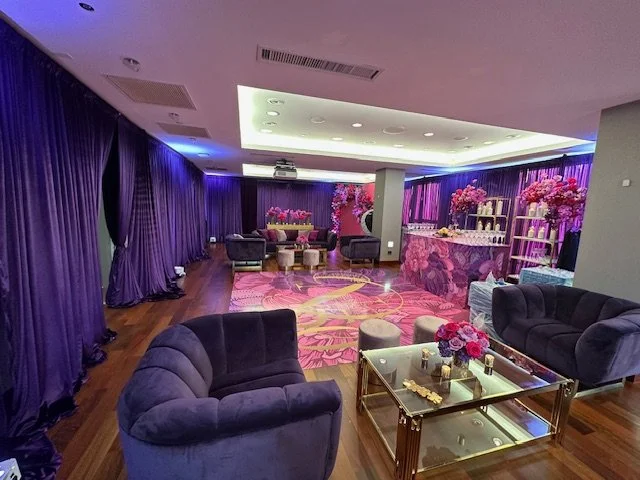 Elegant lounge decorated with purple curtains, velvet sofas, pink flowers, and decorative lighting, featuring a modern, floral-themed setup.