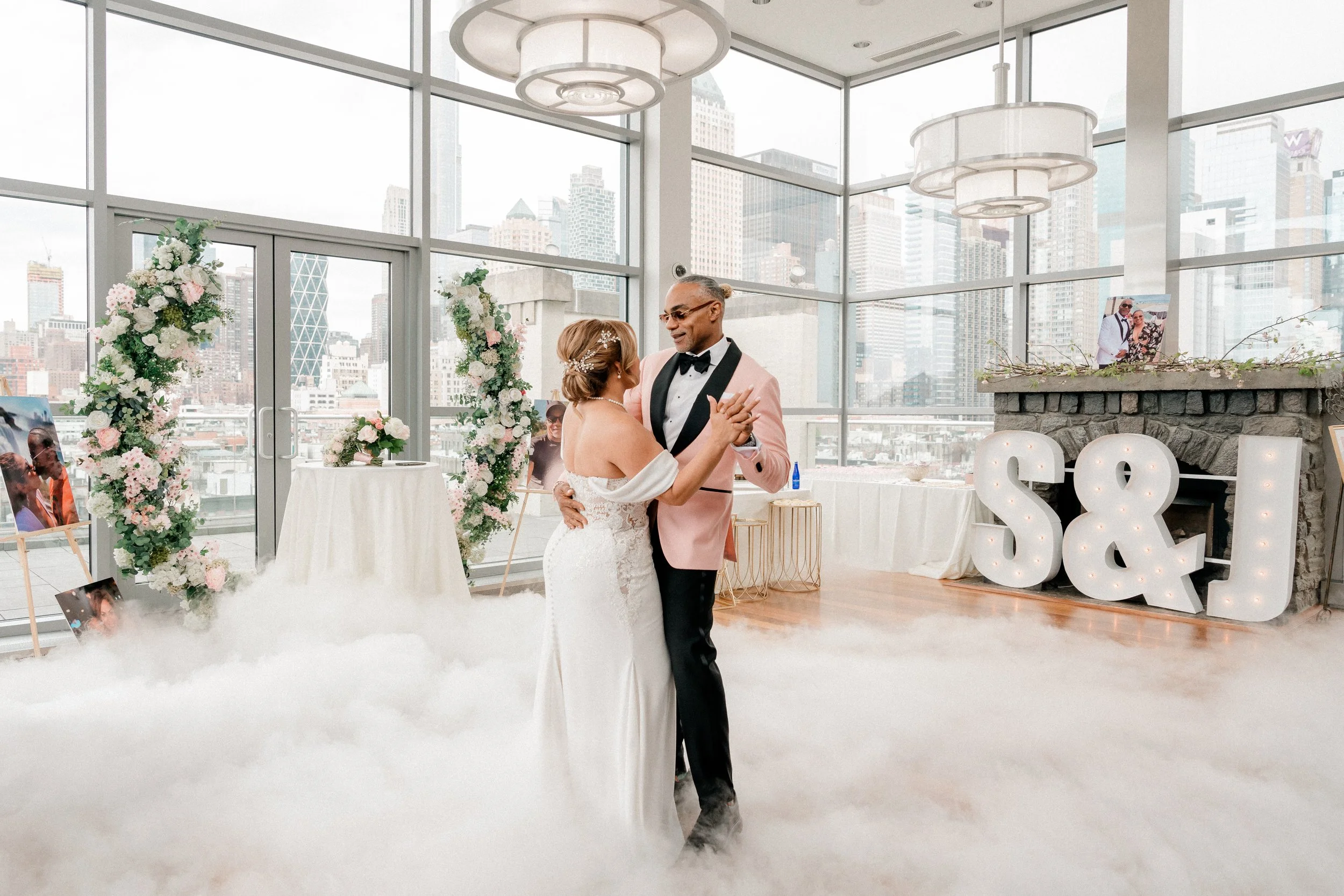 A bride and groom dancing at wedding reception in a bright, modern venue with floor-to-ceiling windows showing a city skyline.