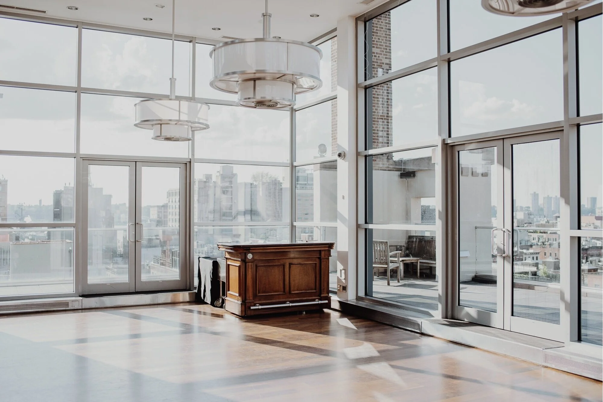 Modern indoor space with large floor-to-ceiling windows, a wooden desk, and two chairs outside on a balcony, overlooking city buildings.