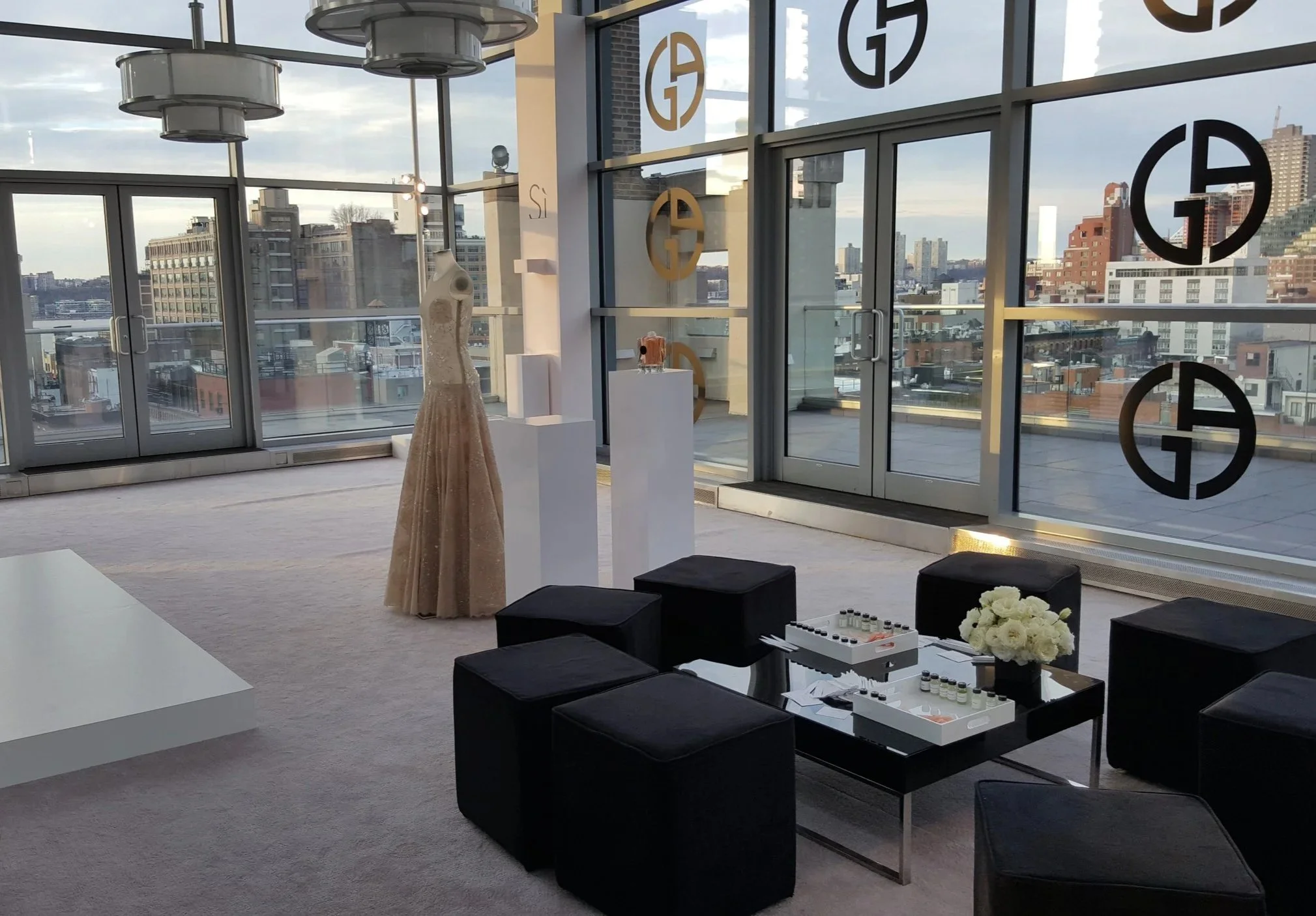 Interior of a modern high-rise apartment or showroom with large floor-to-ceiling windows overlooking a cityscape, featuring display mannequins, black ottomans, a white table with sample bottles, and a bouquet of white flowers.