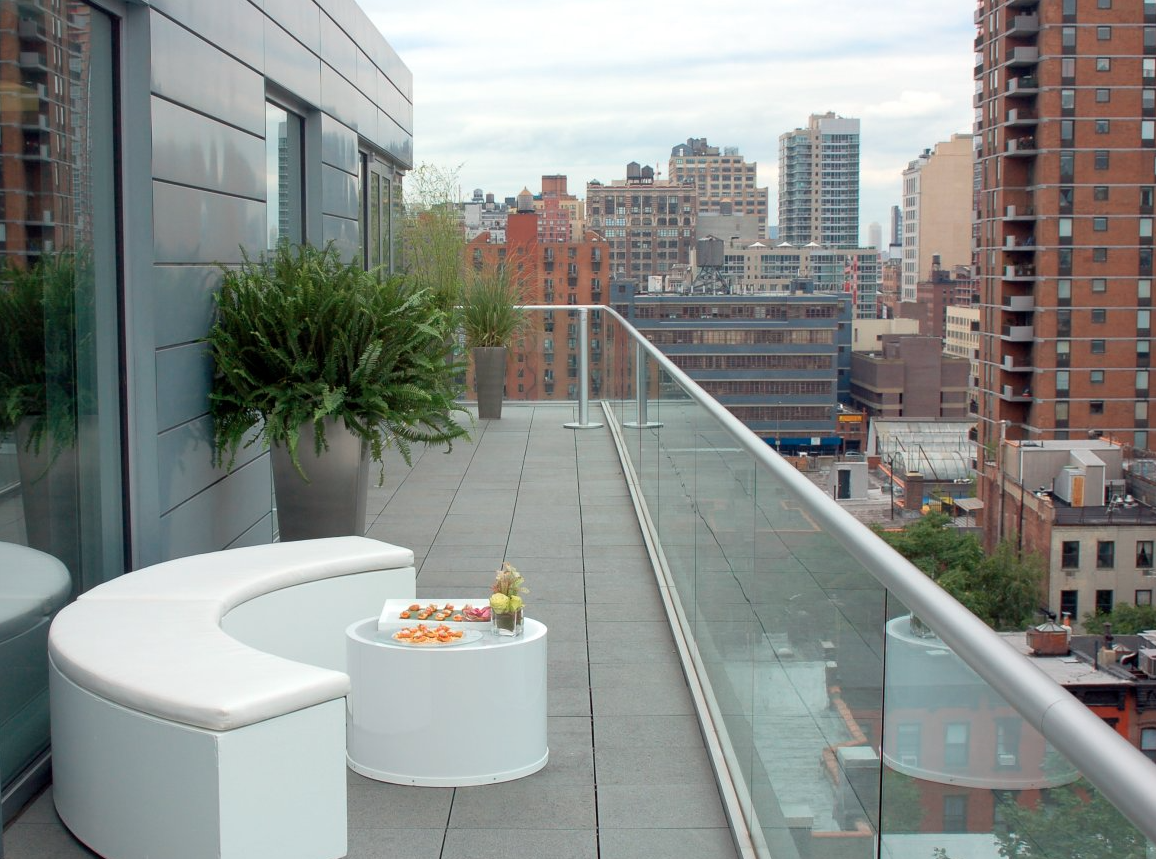 City skyline view from a rooftop balcony with potted plants, a curved white bench, and a small round table with snacks and a drink.
