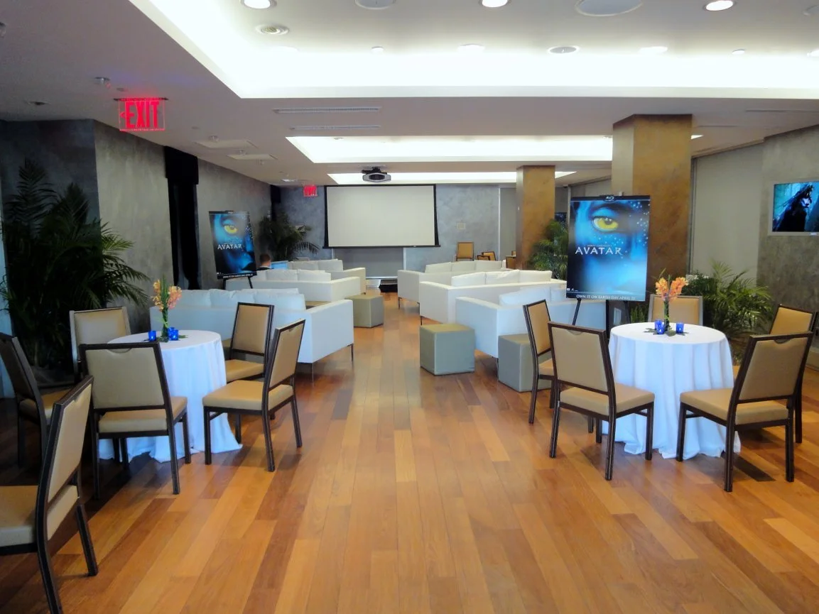 Empty event or conference room set up with round tables and chairs, white armchairs, a large screen at the front, TV monitors, and promotional stands for the movie Avatar.