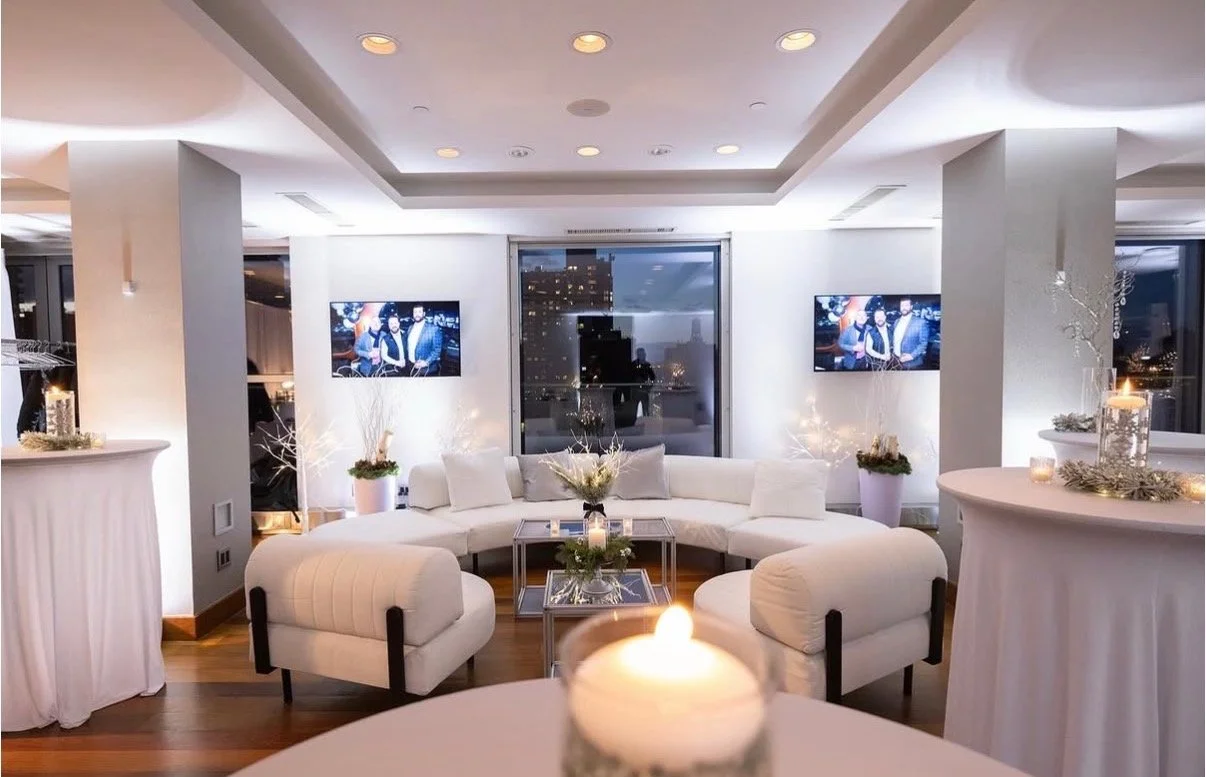 Luxurious modern living room decorated for a festive occasion with white furniture, candles, and string lights, overlooking a city skyline through large windows.
