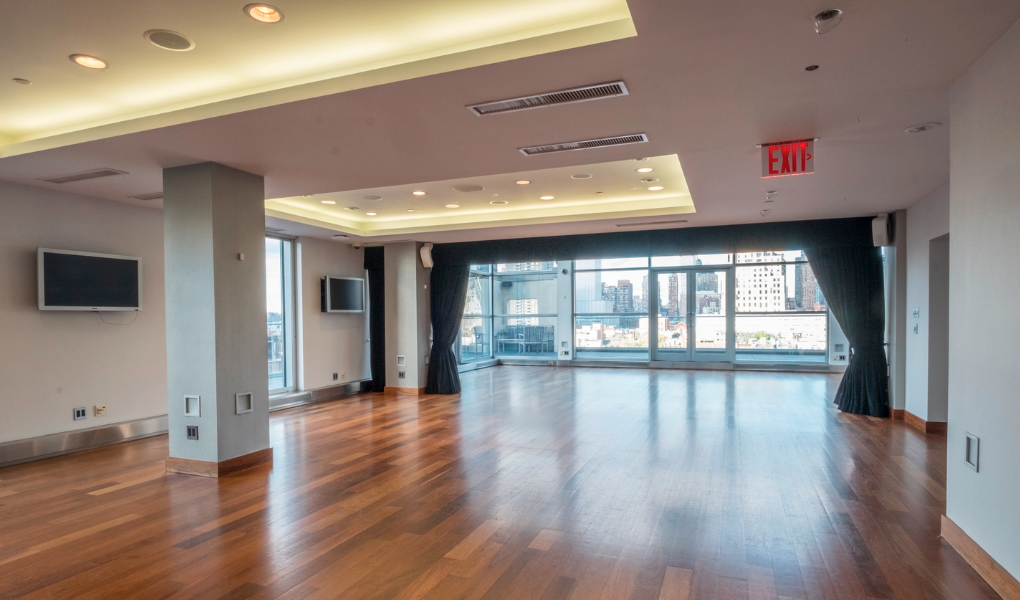 Large, empty room with hardwood floors, two wall-mounted TVs, large windows with city views, dark curtains, ceiling lights, and an illuminated ceiling recess, with an exit sign on the ceiling.