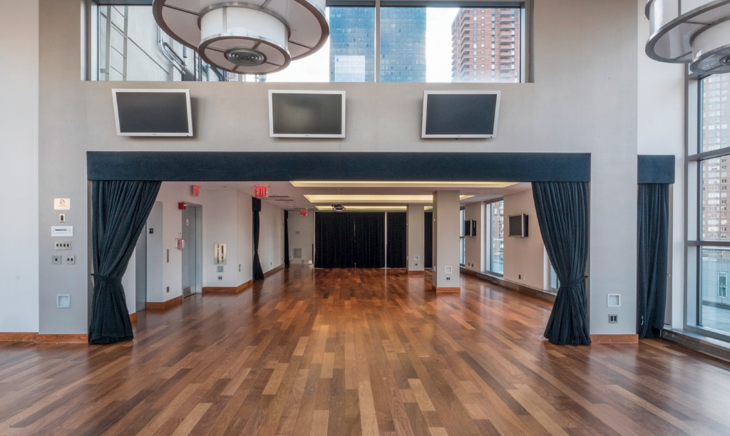 Empty room with wooden floors, large windows, and multiple wall-mounted TVs, with black curtains and city view outside.