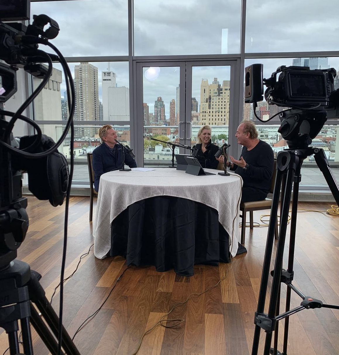 Three people sitting at a round table with microphones, recording a podcast or interview. Large windows in the background reveal a city skyline with tall buildings and cloudy sky.