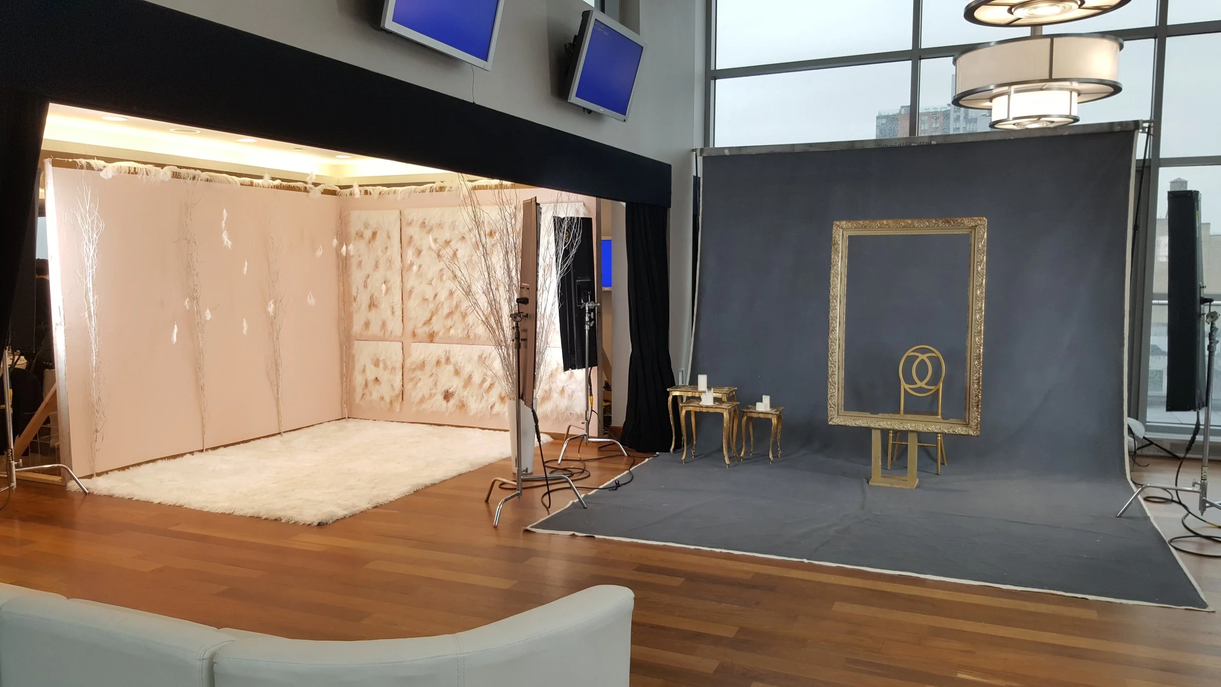 Photo of an indoor photo studio set with a pink tufted backdrop, white rug, gold side tables with candles, a gray backdrop with a gold frame and decorative art, and professional photography lighting equipment. Large windows in the background show cit