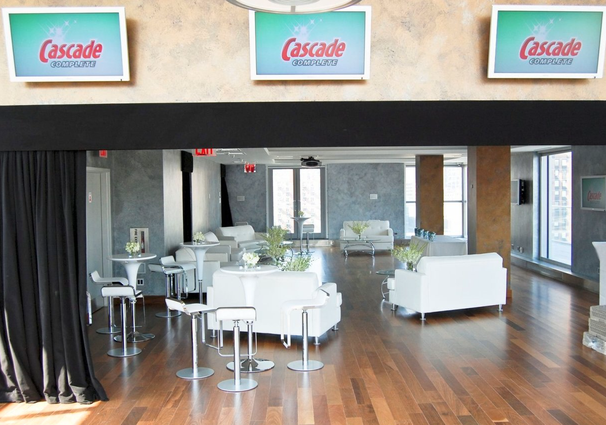 A modern lounge area with white sofas, small tables with floral arrangements, and high chairs, illuminated by large windows, with three screens displaying the Cascade Complete brand logo hanging on the wall.