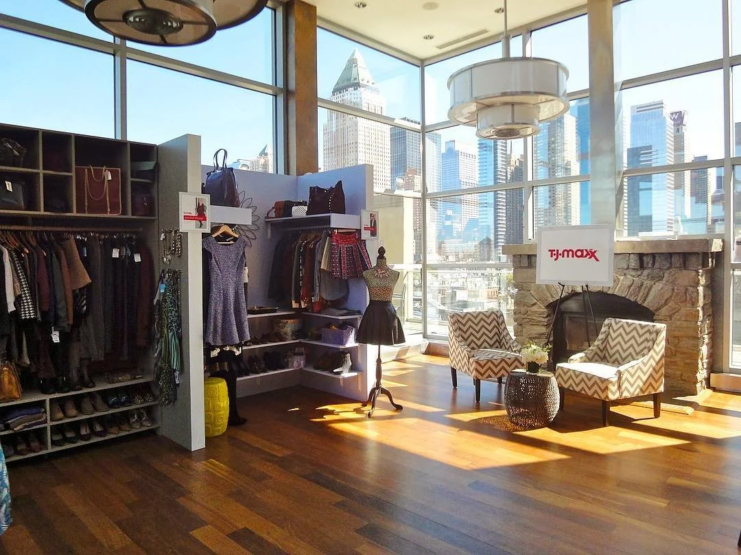 A boutique clothing store with large windows showing a city skyline. The store has shelves of shoes, mannequins dressed in outfits, and seating area with two patterned chairs and a small table with flowers.