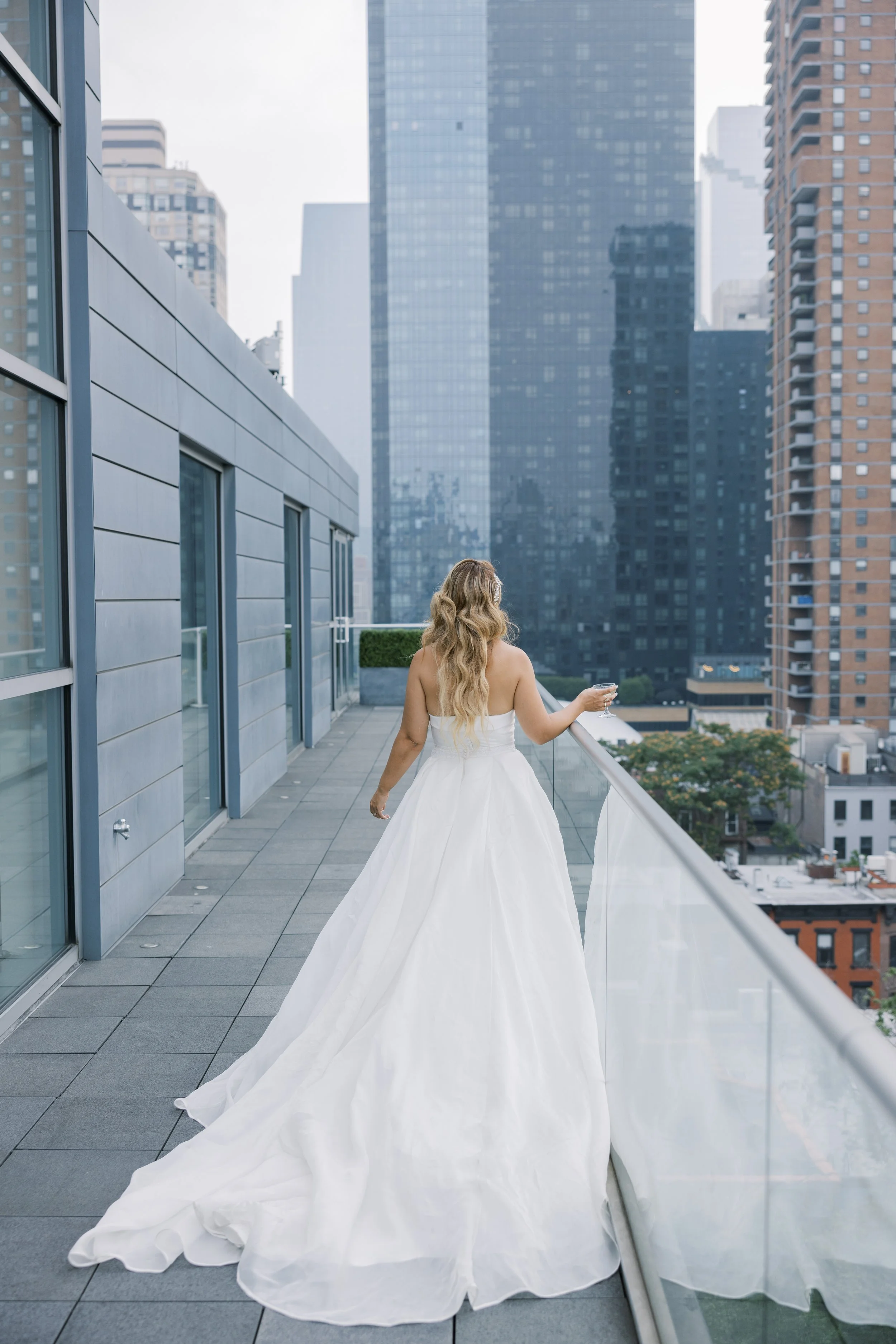 A woman in a white wedding dress on a balcony overlooking a city skyline with tall buildings.