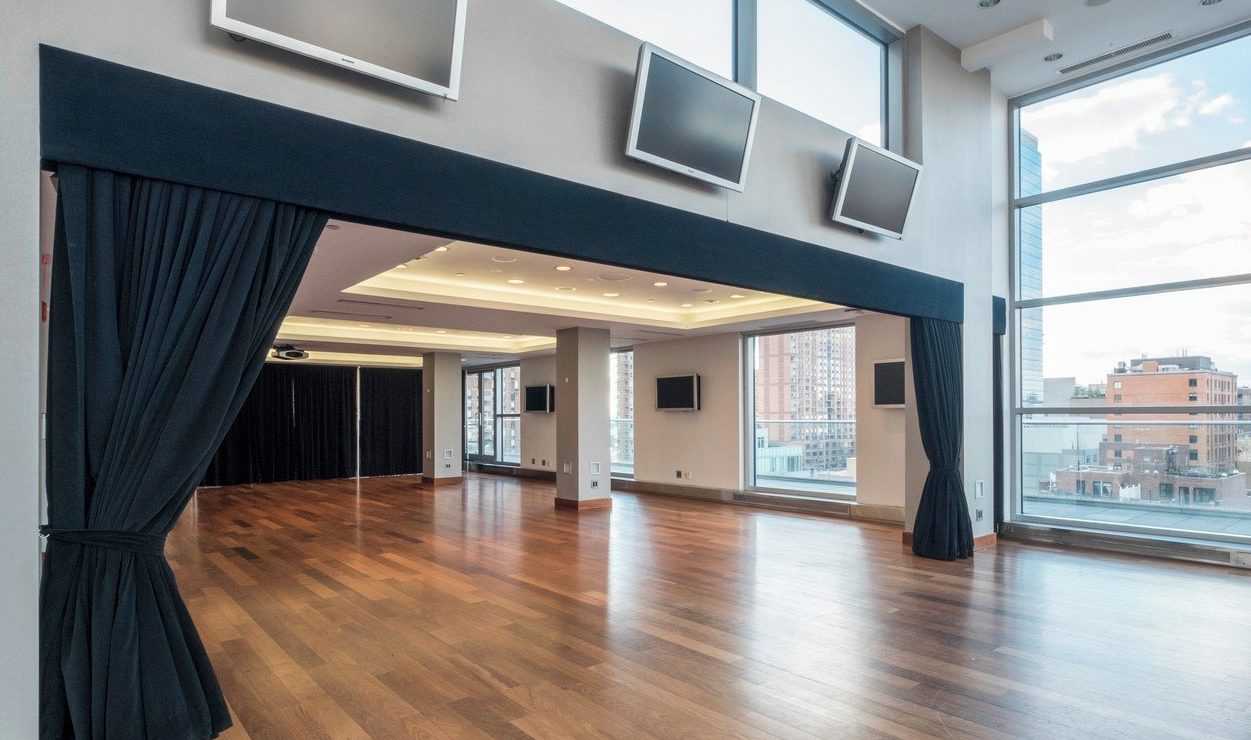 Empty modern room with wooden floors, large windows, multiple wall-mounted TVs, and black curtains.