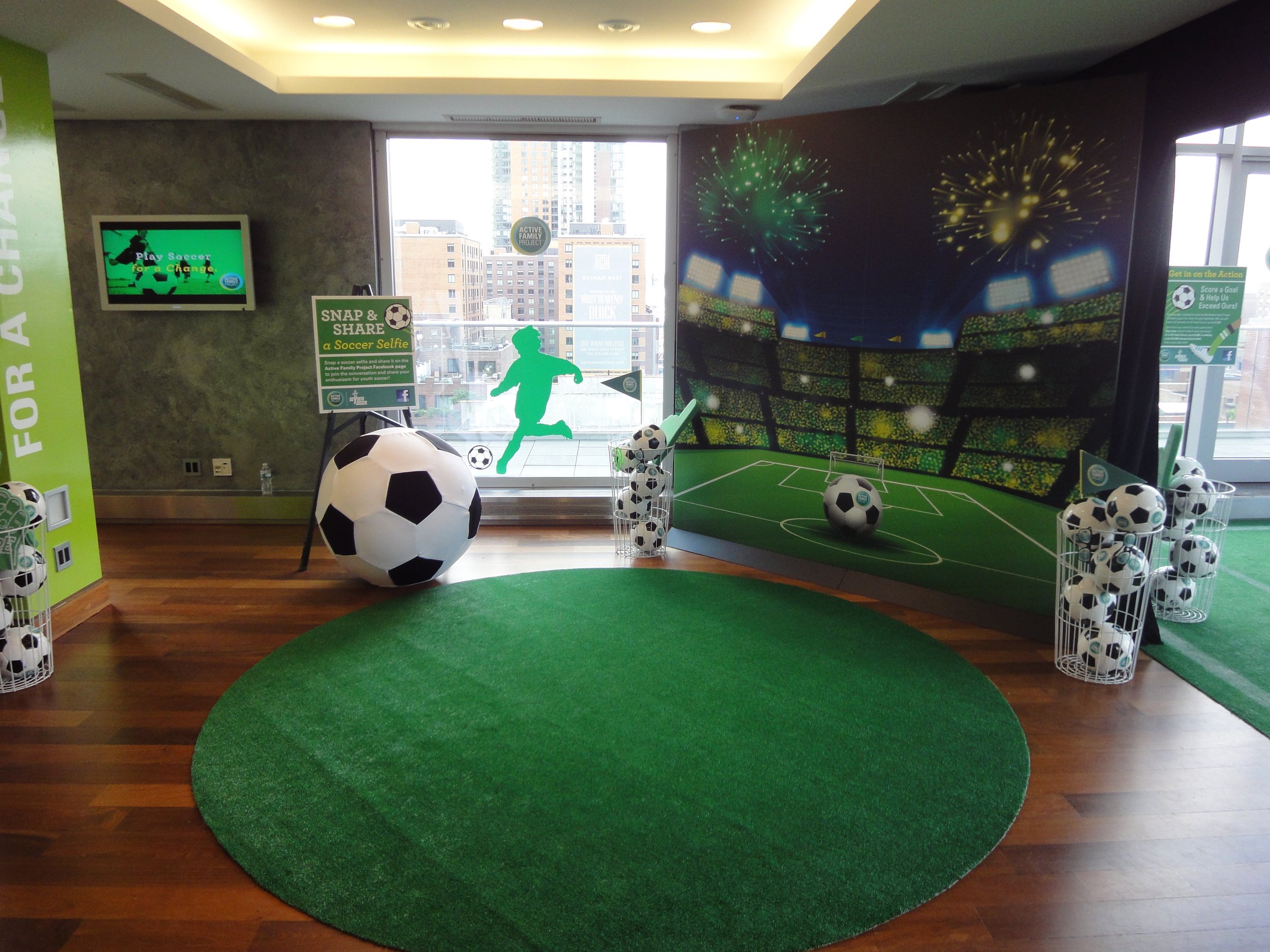 Indoor soccer-themed display area with a mini soccer field, soccer ball props, and bright wall graphics of fireworks and stadium seating, near a large window.