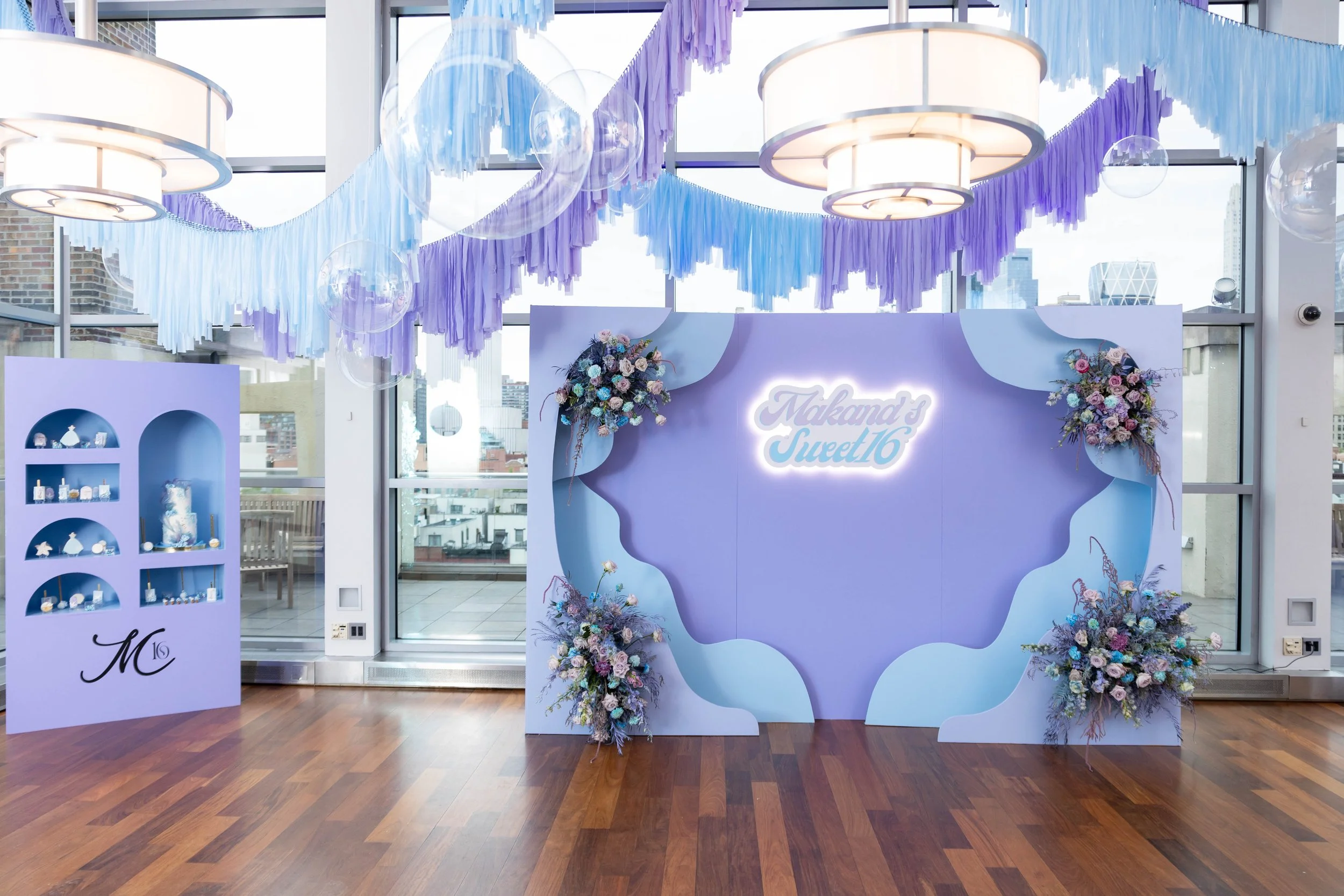 Decorative backdrop at an indoor event with purple and pastel blue fringed hanging decorations, floral arrangements, and a sign that says 'Makand's Sweet 16'.