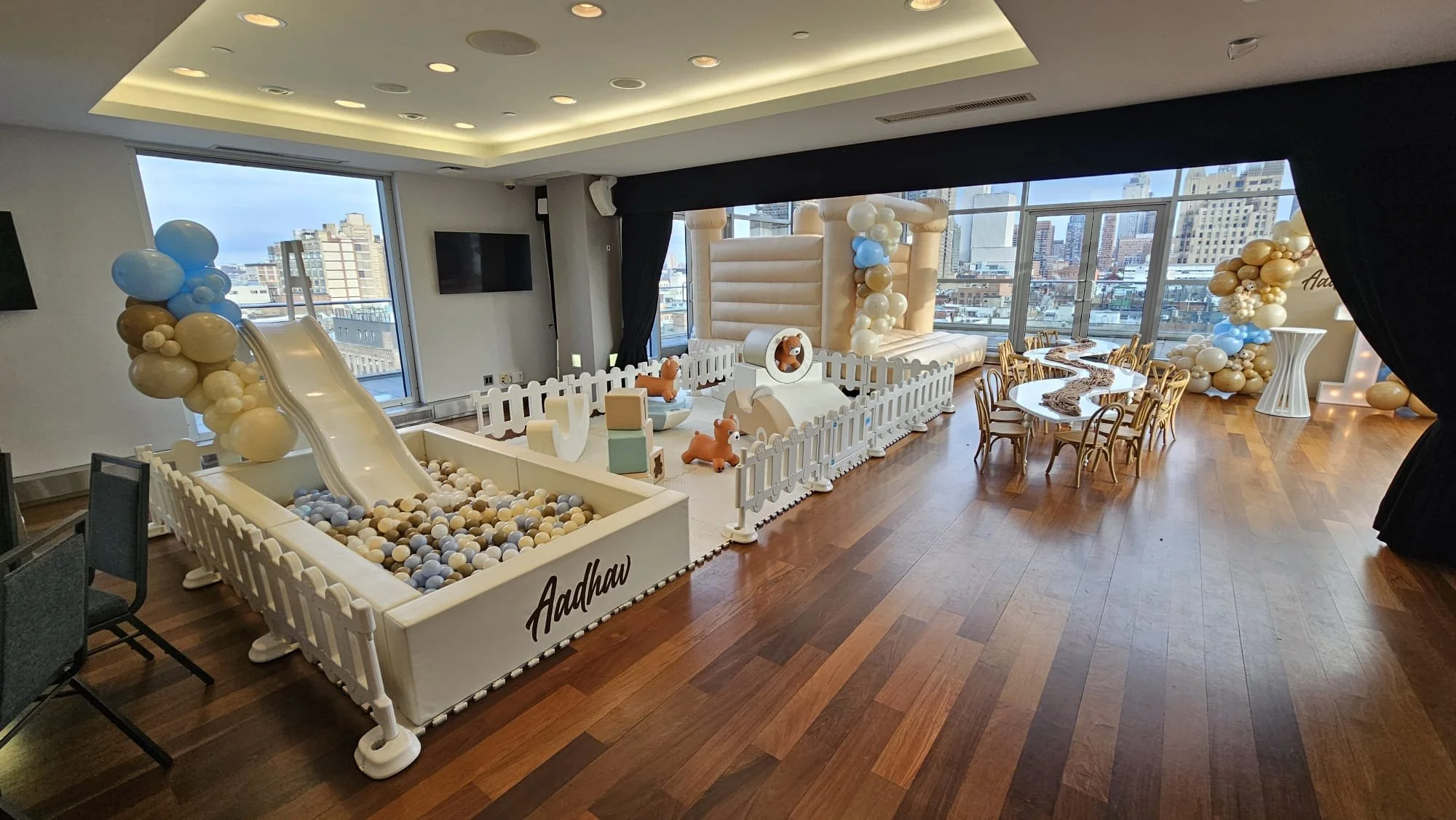 Indoor event space decorated with balloons, a ball pit, small play area, and tables with chairs for children, overlooking city buildings through large windows.