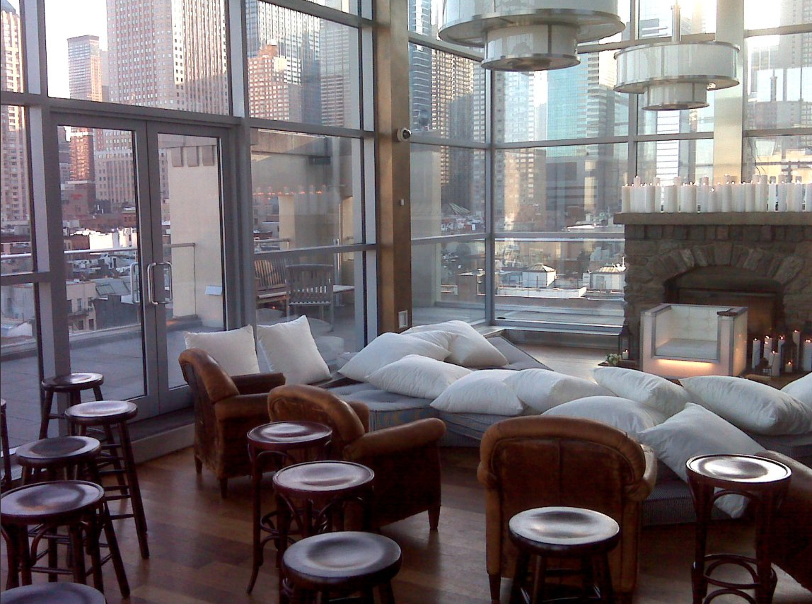 Indoor lounge area with large windows overlooking city buildings, a fireplace with candles, and comfortable seating with cushions and armchairs.
