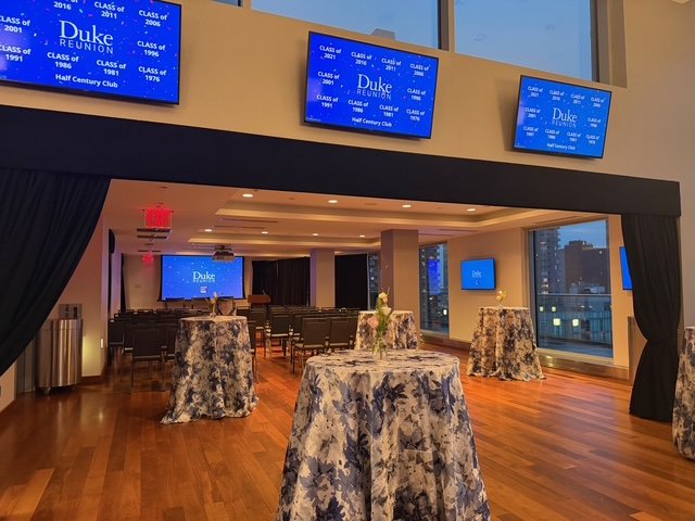 An event space set up for a Duke reunion, with multiple high-top tables covered in floral tablecloths. There are several large screens on the walls displaying reunion information, and windows with a city view. The room has hardwood floors and is deco