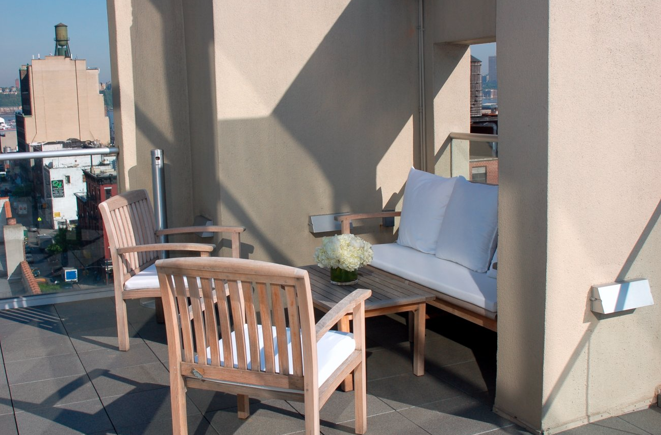 Rooftop patio with wooden chairs, a small table with a flower vase, cushioned bench, and cityscape view with buildings and a water tower.