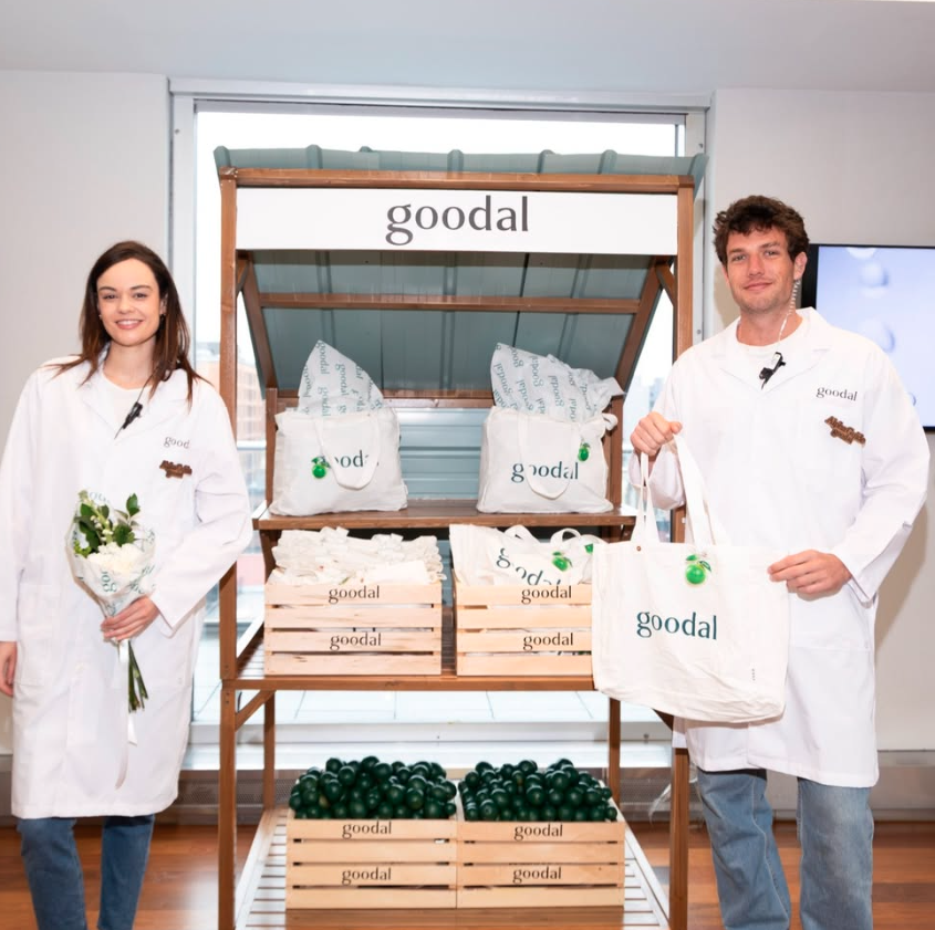 Two people in white coats standing by a display of Goodal-branded products, including wooden crates filled with green balls, paper bags, and boxes, at an indoor event or store.