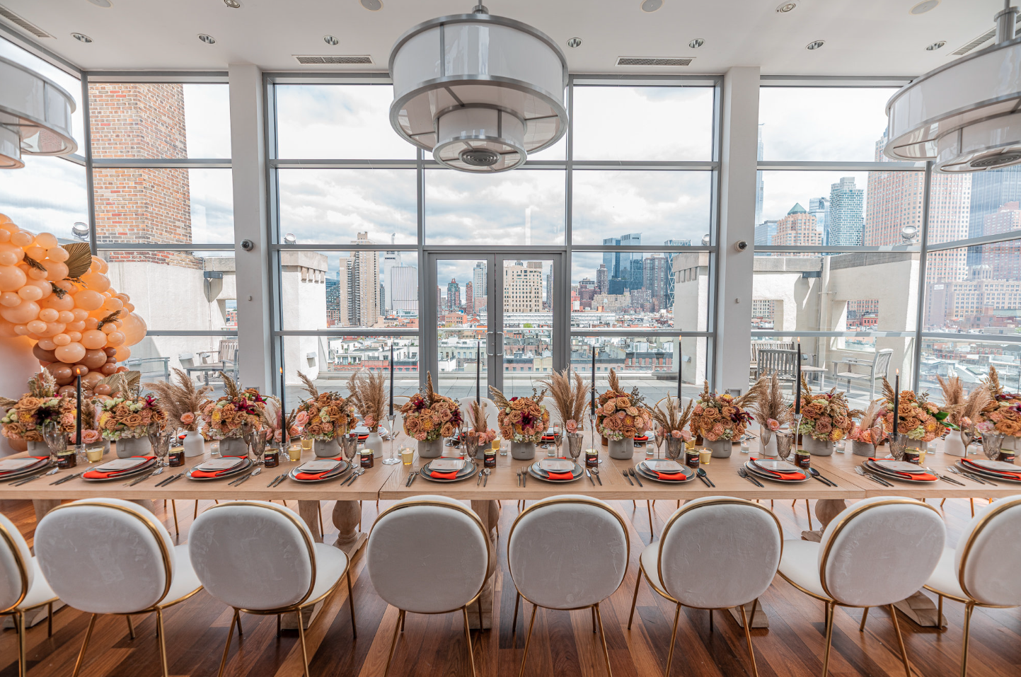 A long dining table set for a celebration with floral centerpieces, placed near large windows with a city skyline view.