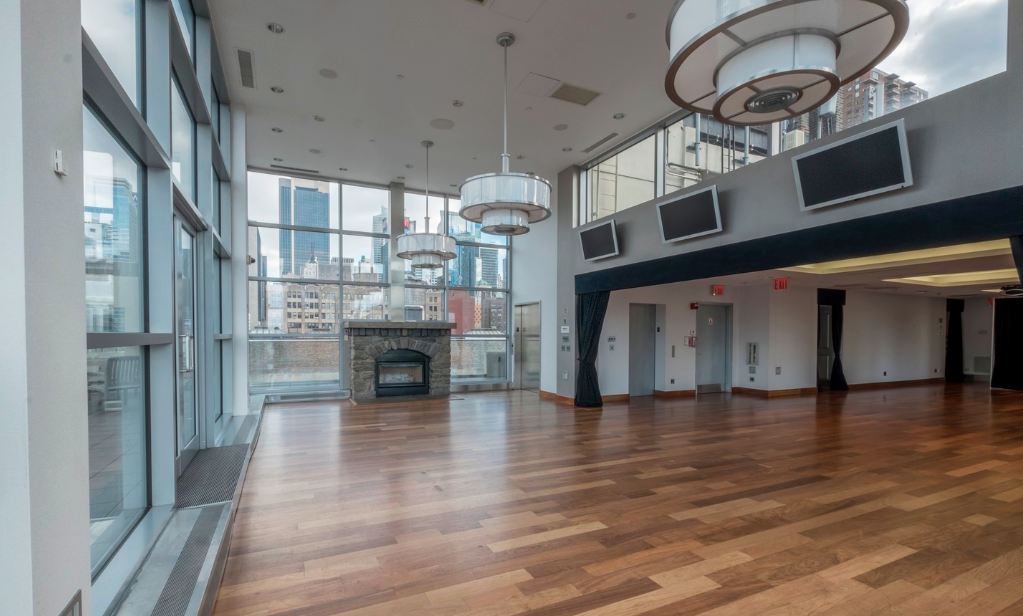 Empty modern event space with large windows, hardwood floors, a stone fireplace, multiple ceiling lights, and several wall-mounted screens.