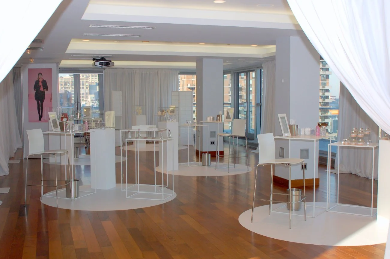 Empty event space with white display tables and chairs, set up for a presentation or product showcase, with city view through large windows.