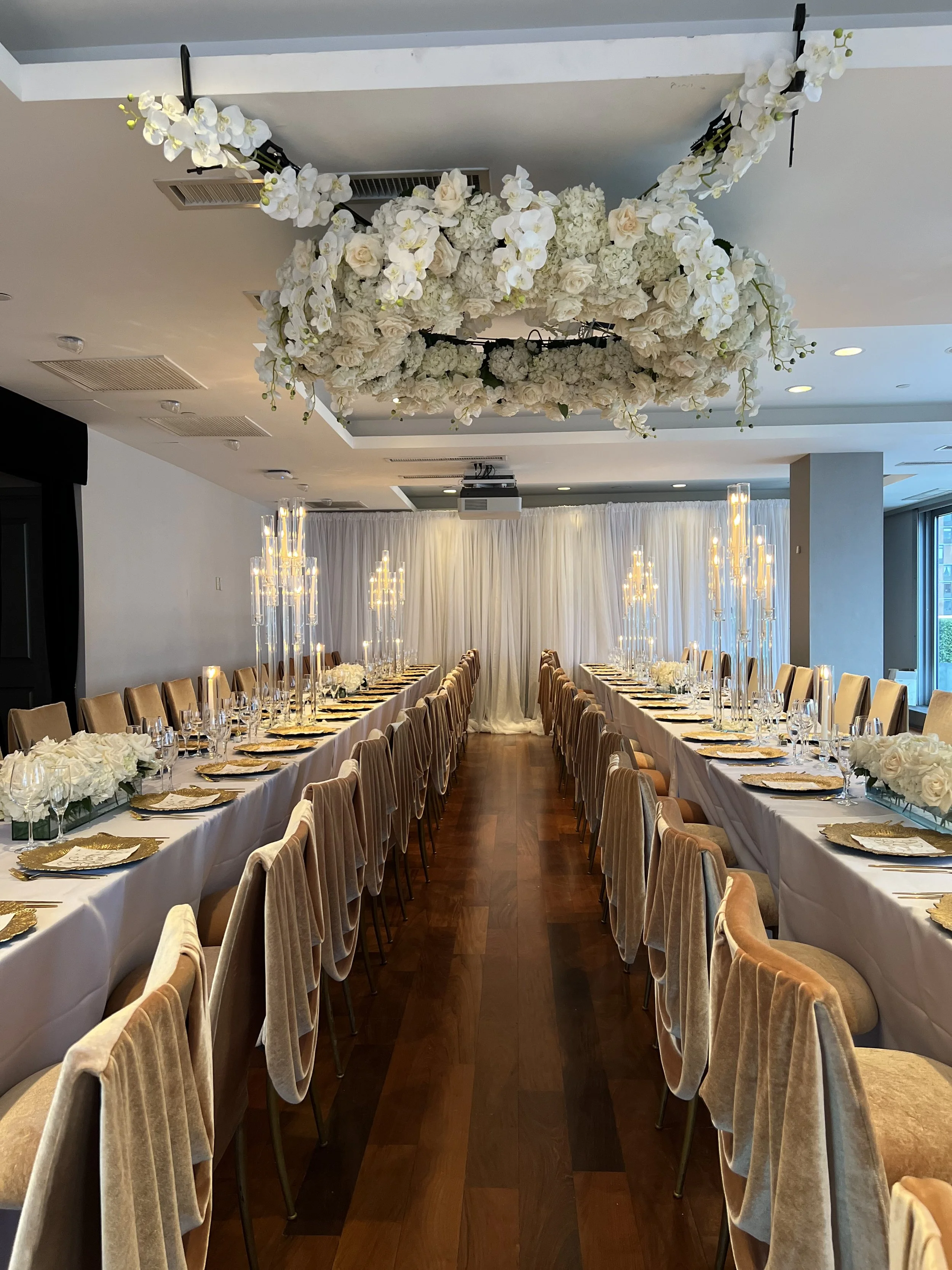 A large banquet hall decorated for a formal event with long dining tables covered in white tablecloths, beige chairs, floral centerpieces, and tall candle holders, with a floral chandelier hanging from the ceiling and a white curtain backdrop at the 