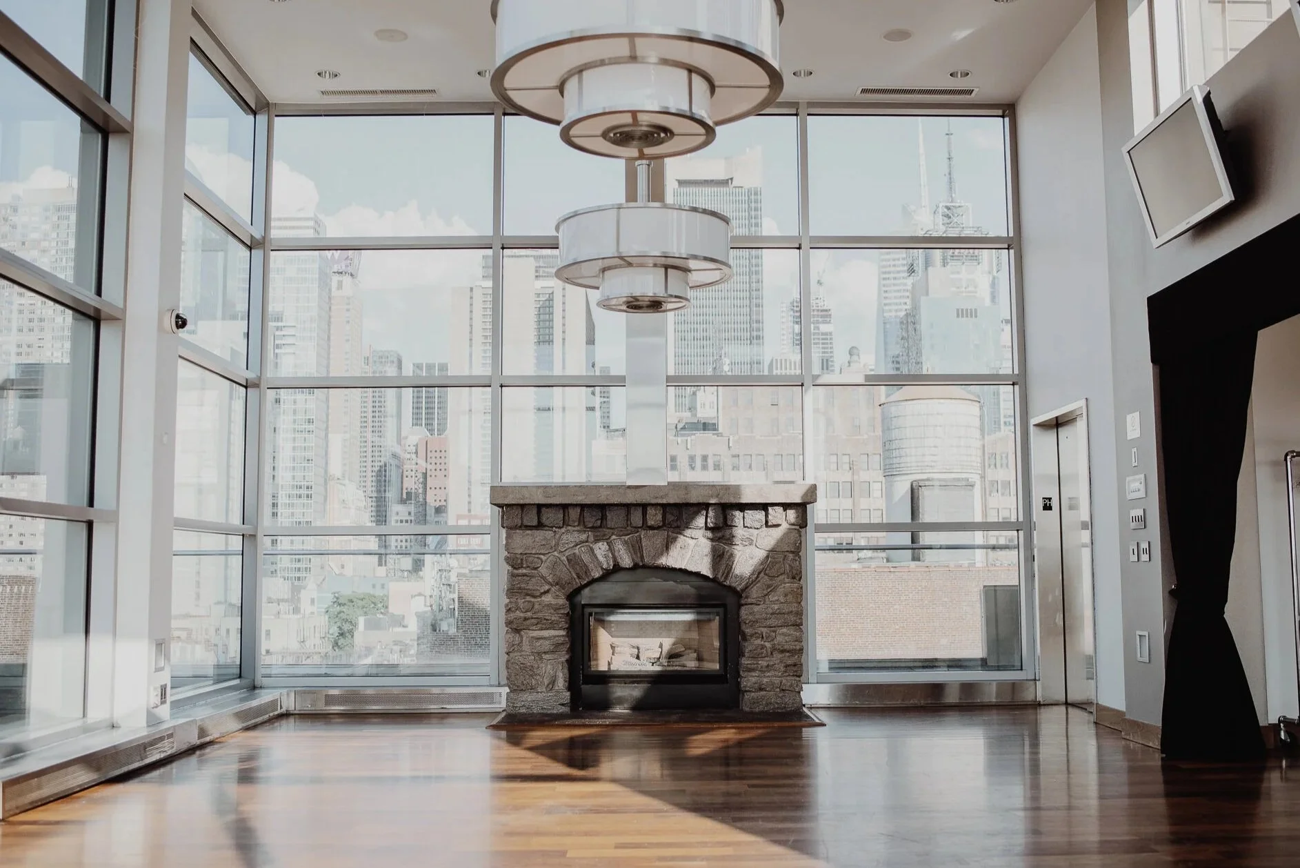 Interior view of a modern city apartment or hotel lobby with large floor-to-ceiling windows showing a cityscape of tall buildings. There is a stone fireplace in the center, a large round chandelier hanging from the ceiling, and a wall-mounted TV on t
