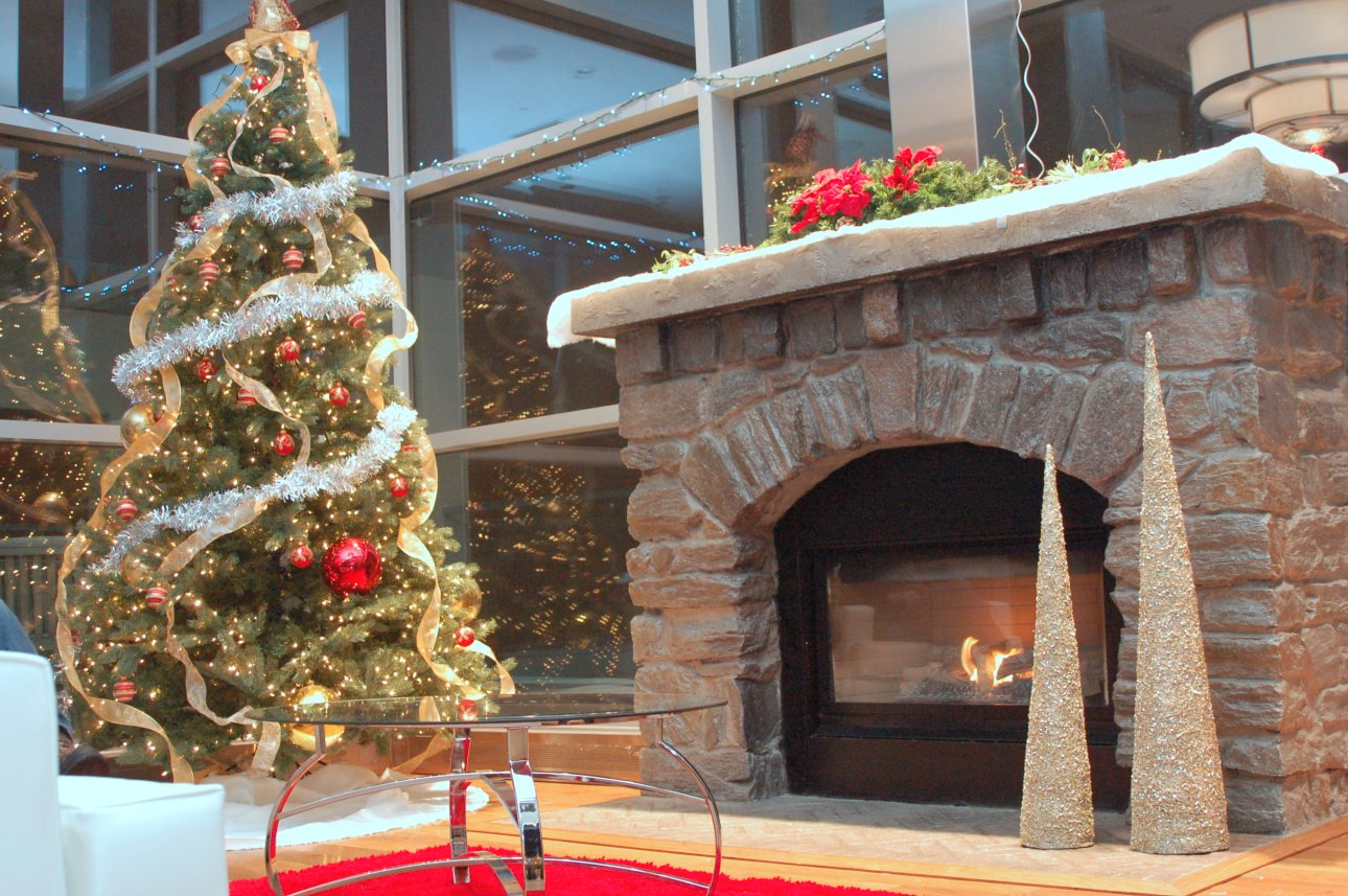 Decorated Christmas tree with gold and red ornaments, tinsel, and ribbon next to a stone fireplace with a fire burning. Two gold glitter cone Christmas decorations stand beside the fireplace, and holiday lights are reflected in the windows.