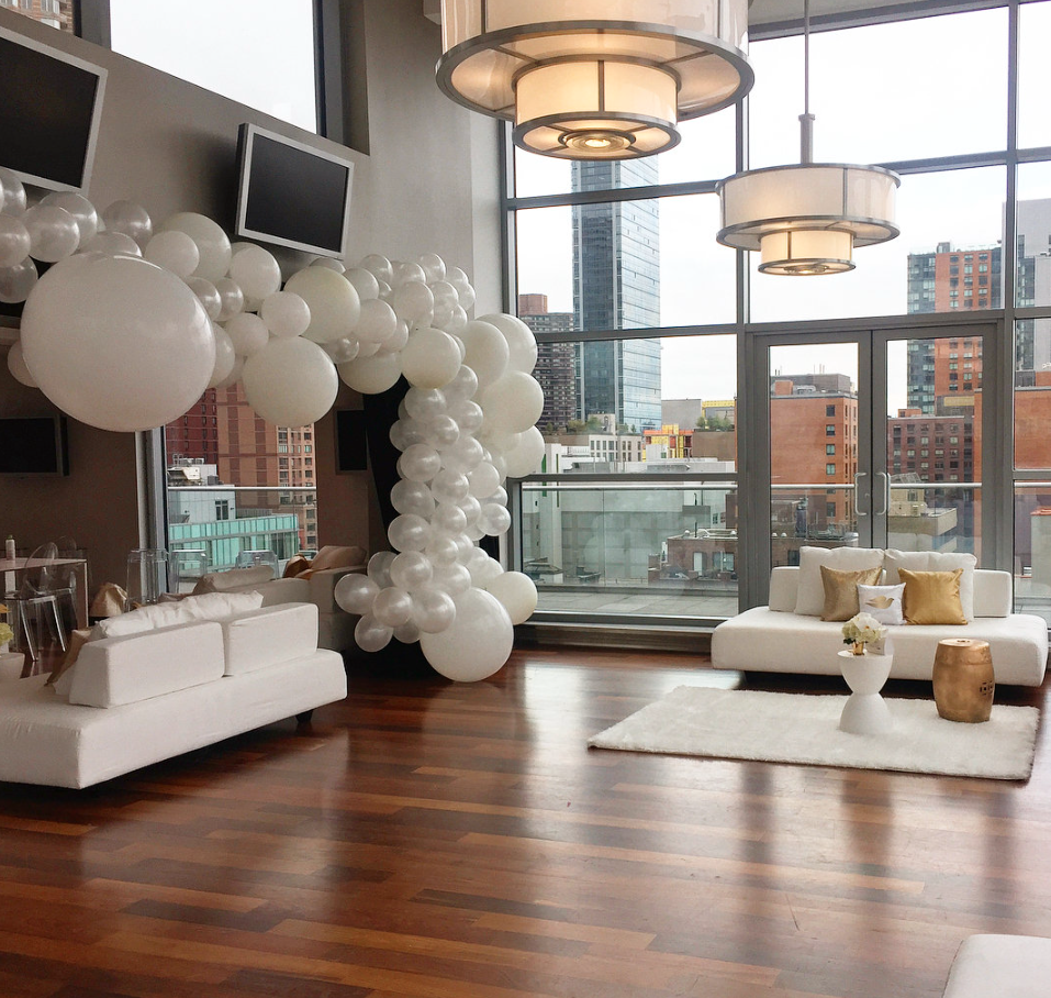 Modern living room with large windows, white couches, a white rug, decorative pillows, and an arch of white balloons.