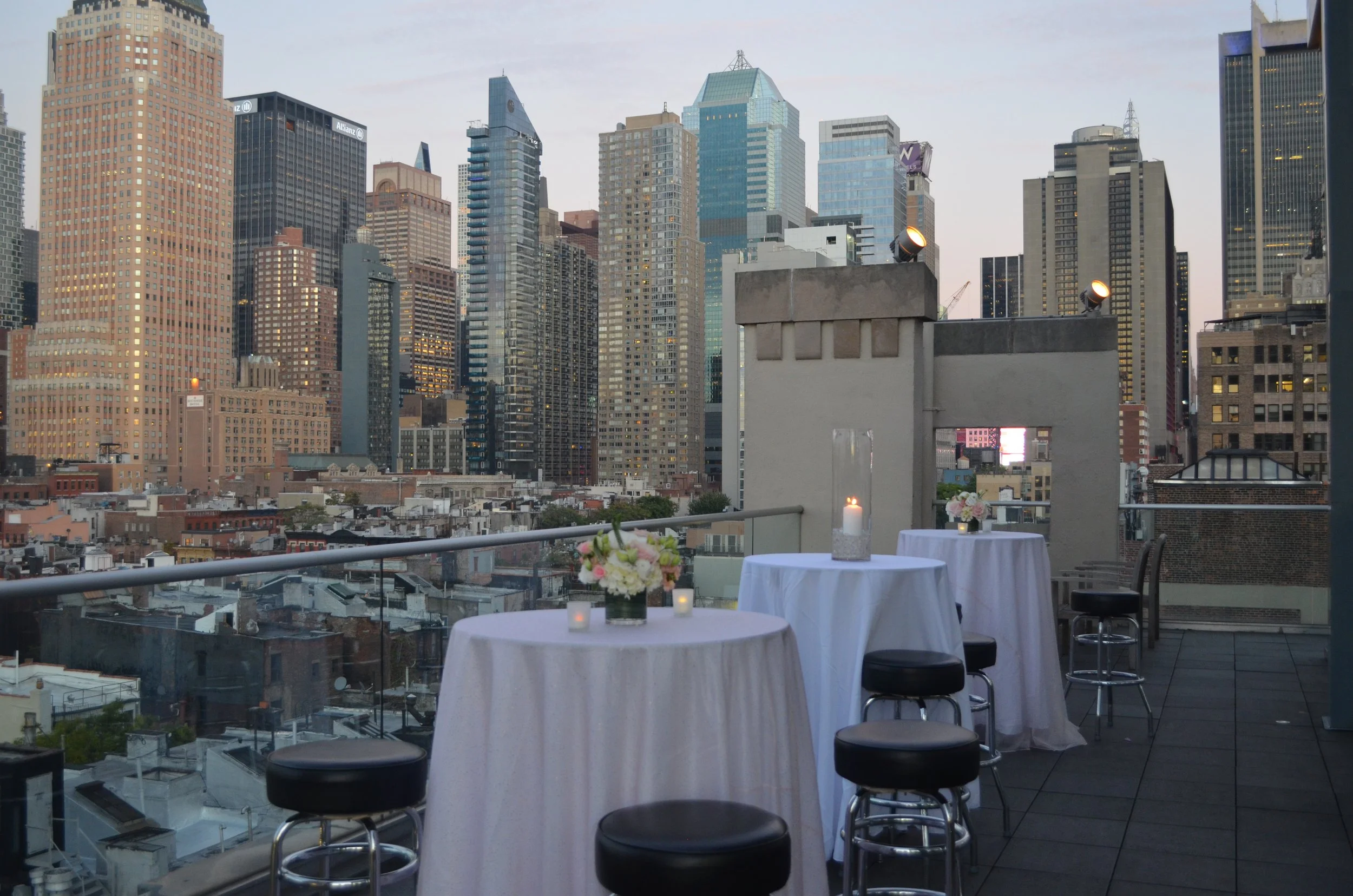 View of a city skyline with tall skyscrapers from a rooftop terrace, featuring tables with white tablecloths, flower arrangements, and candles.
