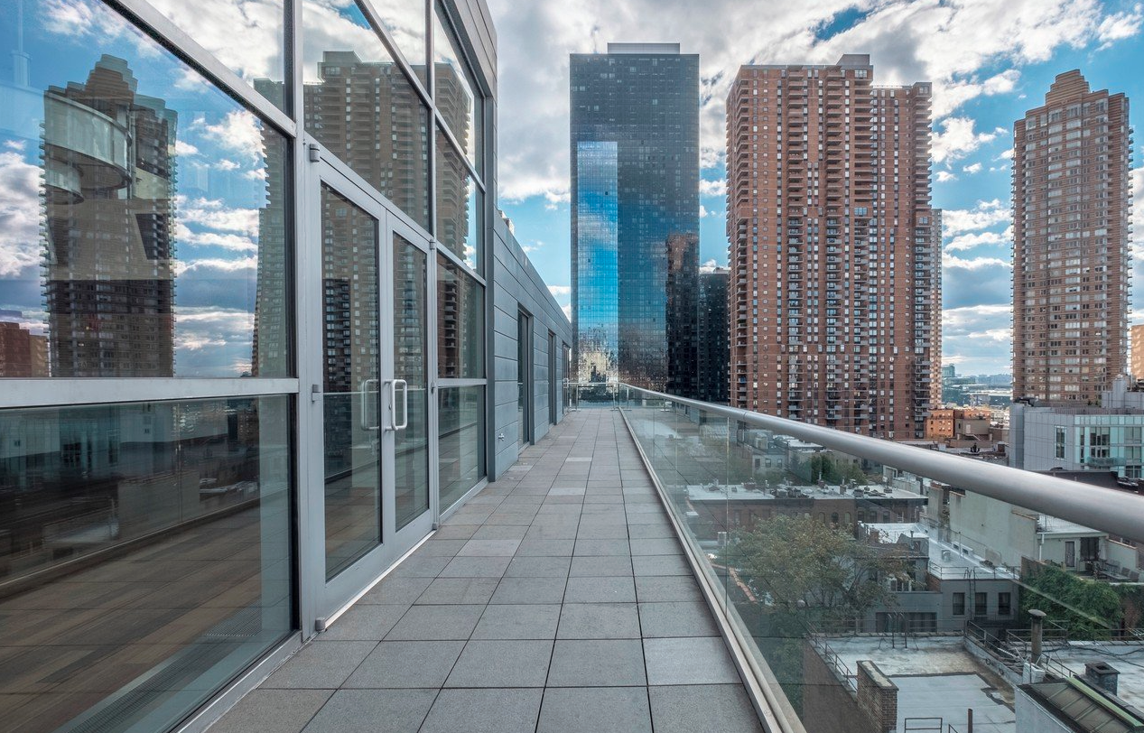 Empty rooftop balcony with glass railing, view of tall city buildings, partly cloudy sky.