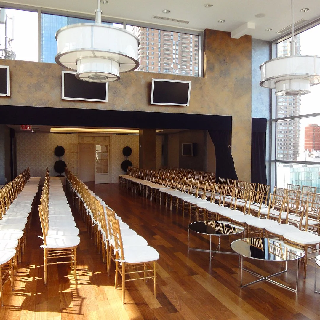 Empty banquet room with rows of gold chairs with white cushions, large windows, modern hanging lights, and wall-mounted TVs.