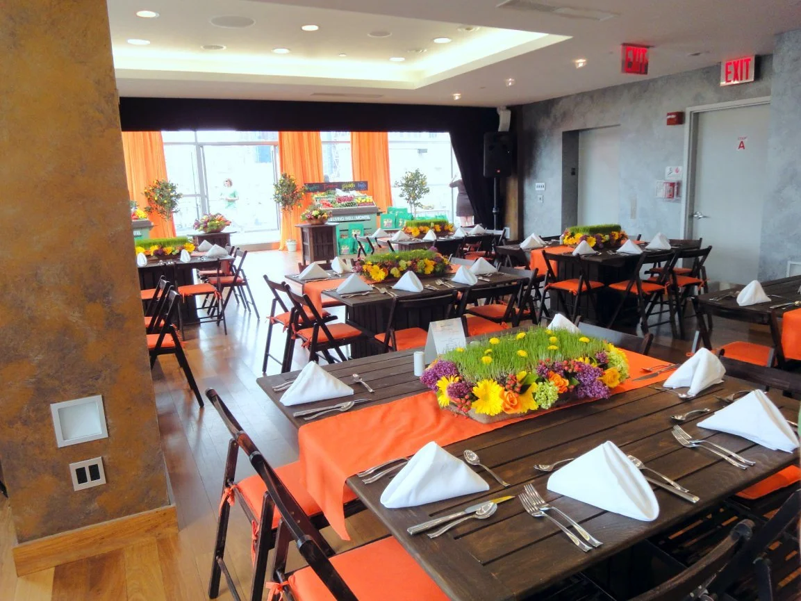Indoor event space decorated with tables covered in orange tablecloths, white napkins, and floral centerpieces, with a stage and large windows in the background.