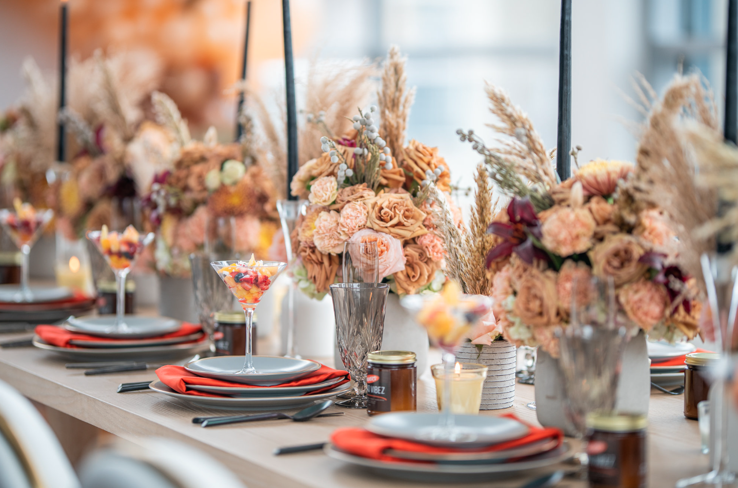 Elegant dining table decorated with large bouquets of pastel-colored flowers, candles, and place settings with gray plates, black utensils, and orange napkins. Glassware and small jars are also on the table.