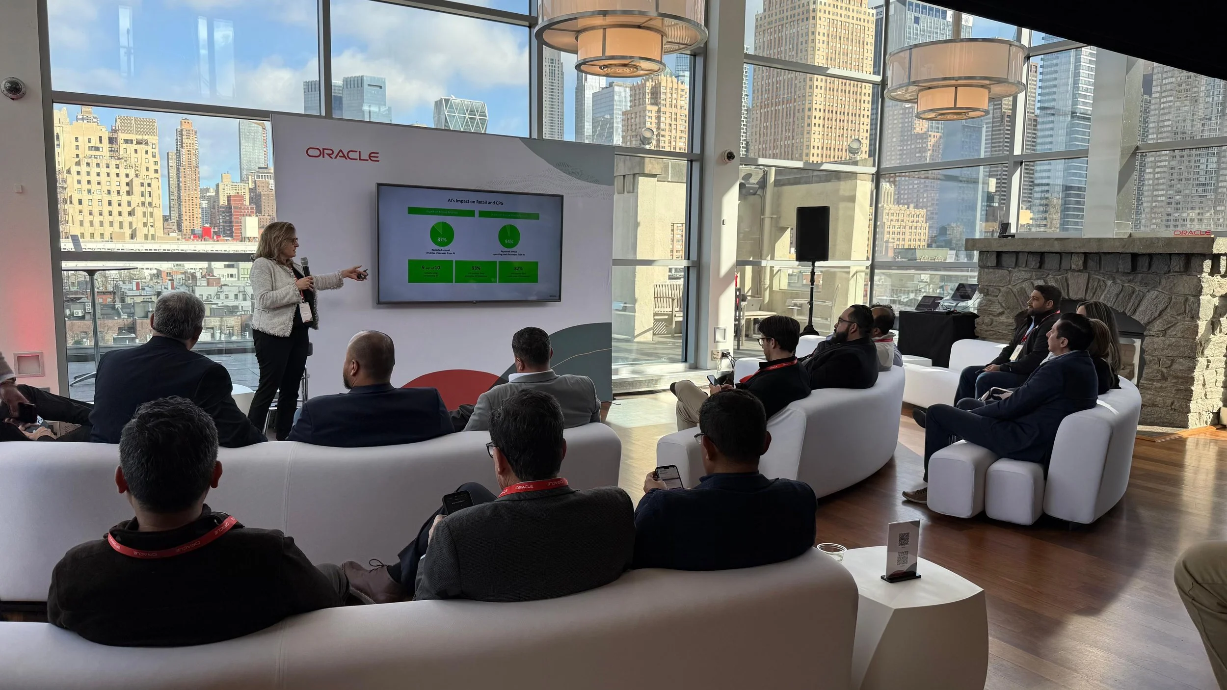 A woman giving a presentation at an event hosted by Oracle in a modern, glass-paneled venue with a cityscape view. Audience members are seated on white sofas, listening and taking notes.