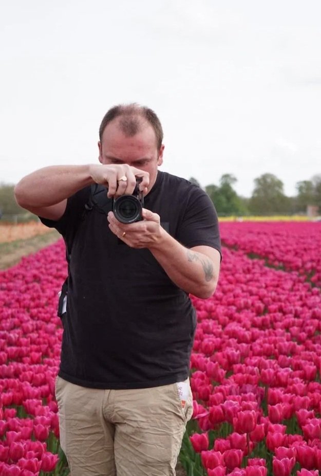 A man in black shirt and khaki shorts taking a photo of himself with a camera in a field of pink tulips.