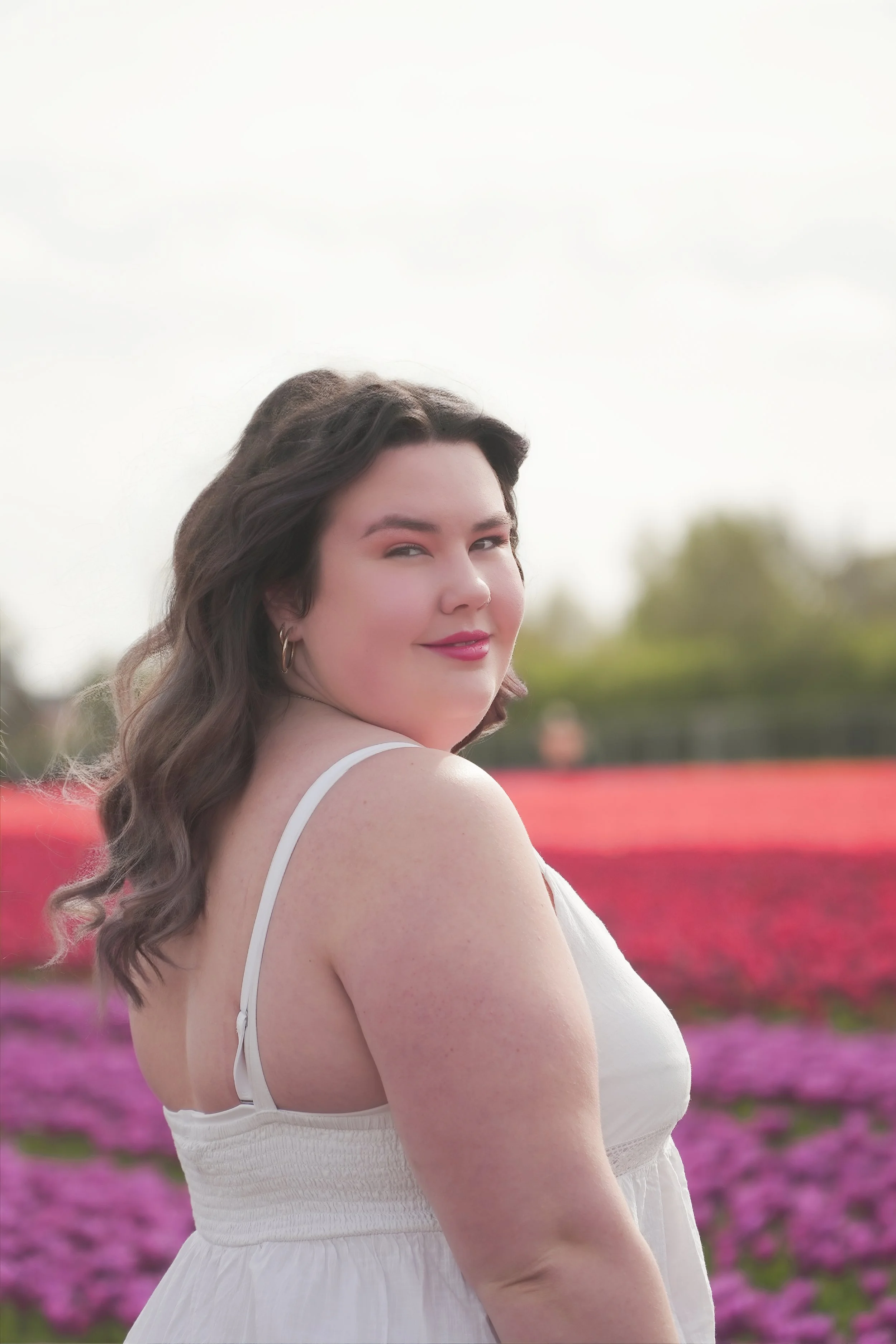 A woman with long brown wavy hair wearing a sleeveless white dress, standing in a colorful flower field with pink and purple blooms, smiling softly at the camera.