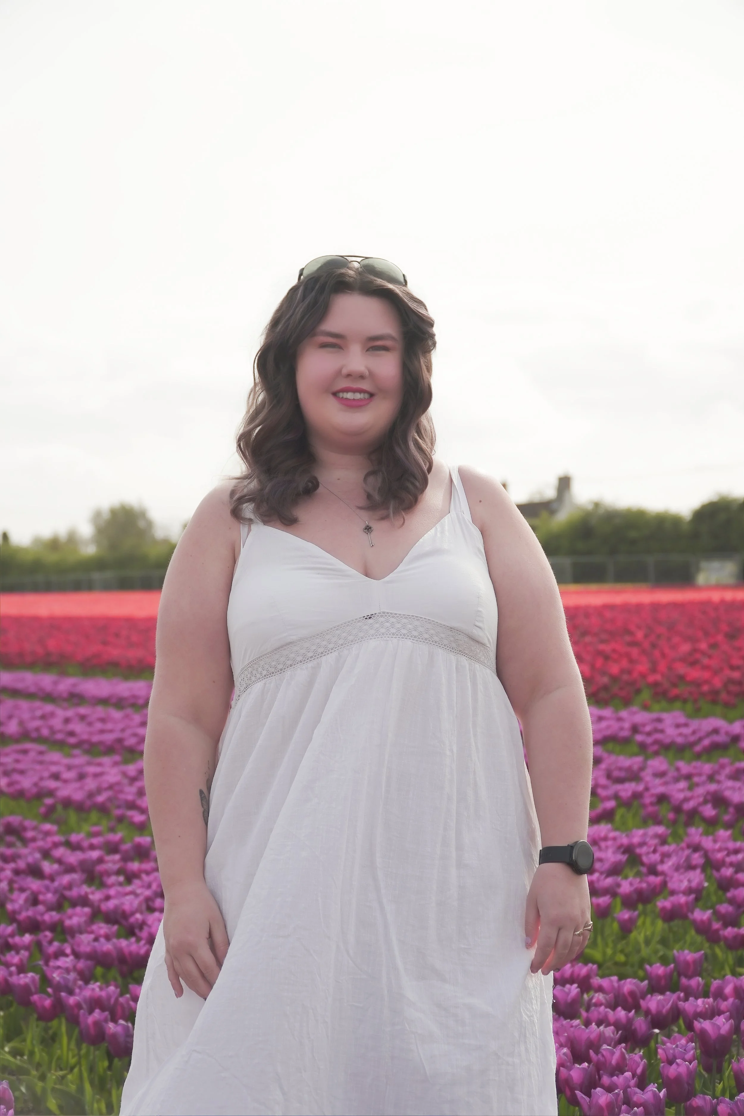 A woman with wavy brown hair and sunglasses resting on her head, wearing a white sleeveless dress, standing in a field of purple and pink flowers with a cloudy sky in the background.