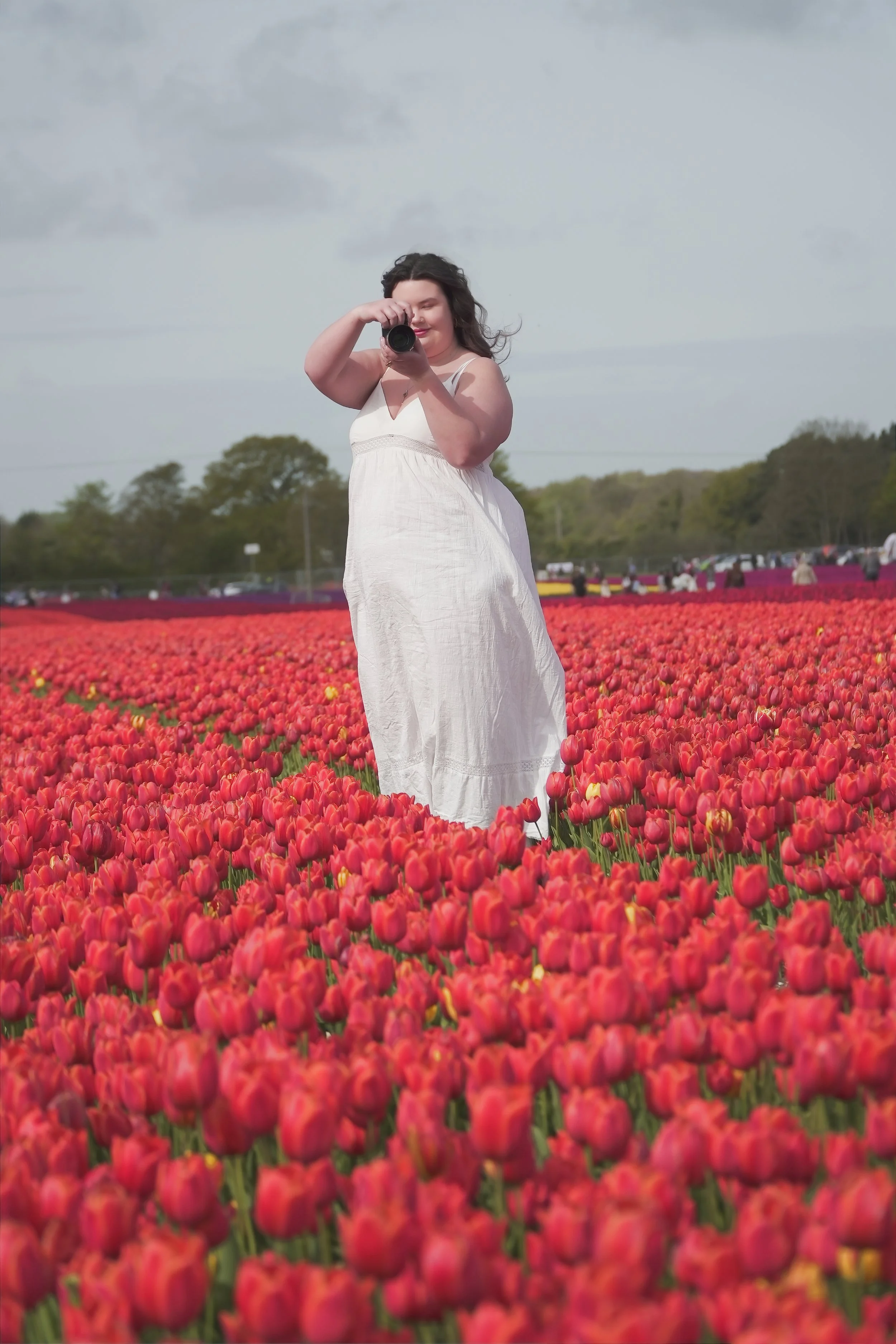 A woman in a white dress standing in a field of red tulips, taking a photo with a camera.