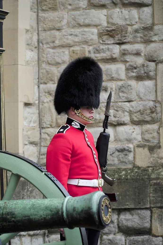 A British royal guard in traditional red uniform with black bearskin hat standing in front of a stone wall, holding a rifle.