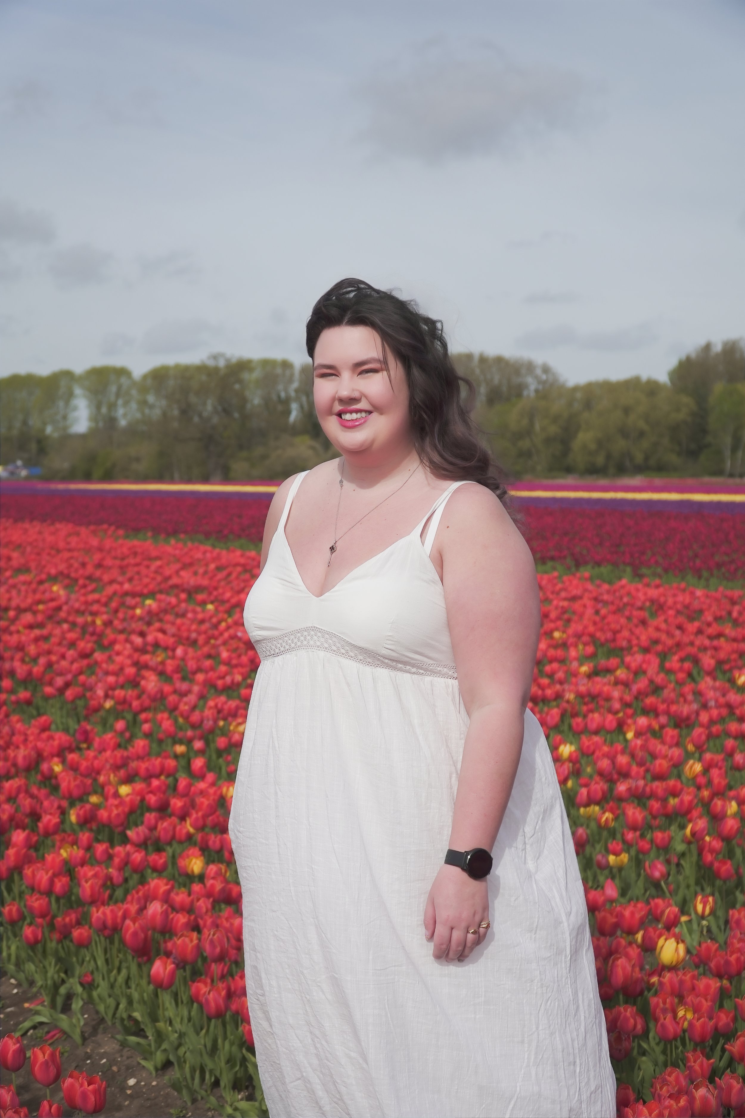 A smiling woman in a white sundress standing in a colorful tulip field with trees and a cloudy sky in the background.
