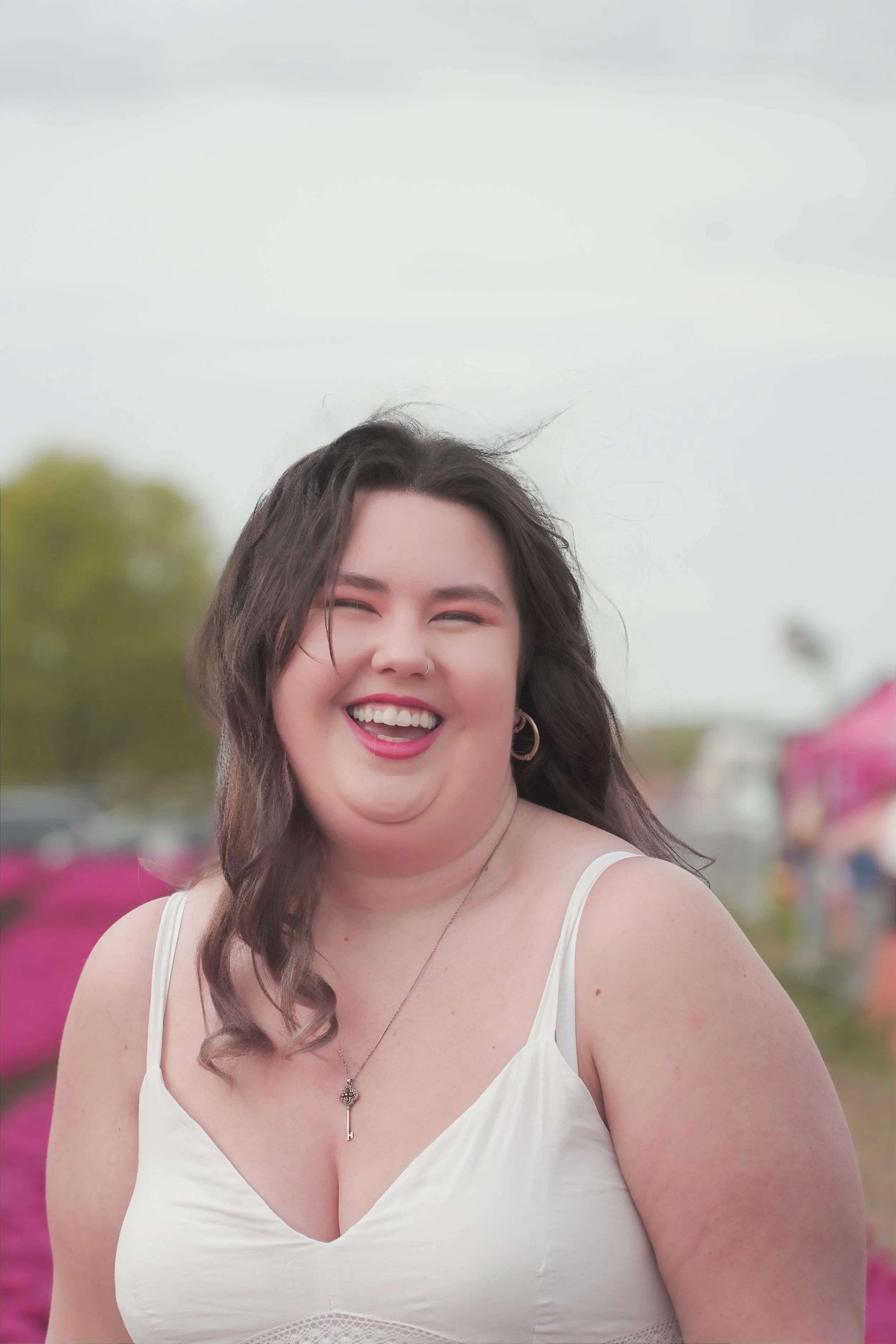 A young woman outdoors, smiling and laughing, wearing a white sleeveless top, with pink flowers in the background.